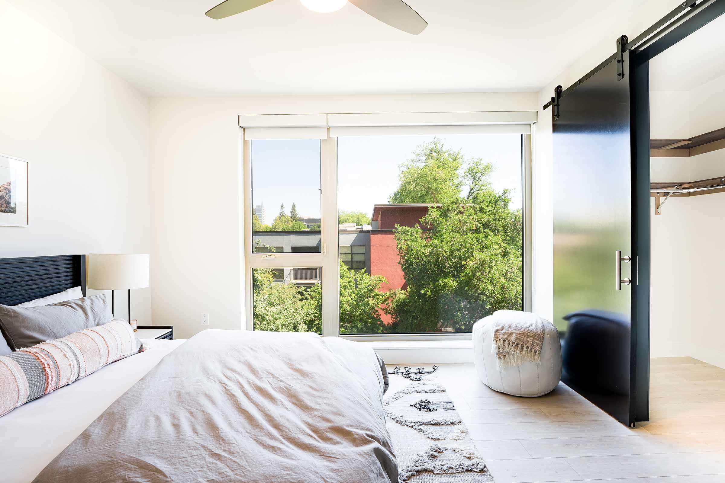 Modern bedroom featuring a large window with a view of trees and a cityscape outside. The room includes a bed with a textured gray blanket, a nightstand with a lamp, and a white pouf. A sliding black door leads to a closet space, enhancing the minimalist design.