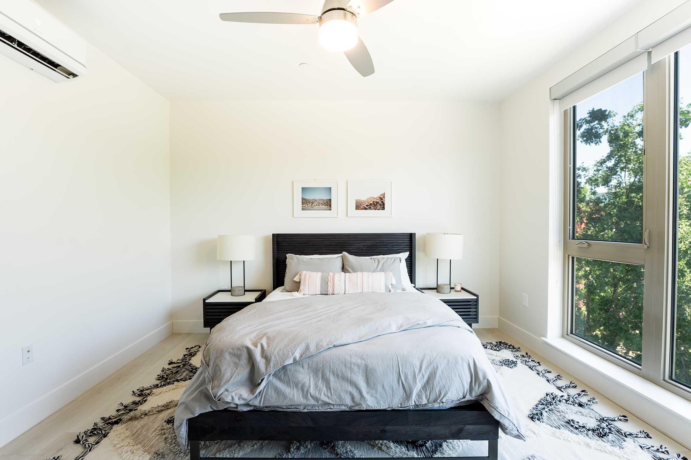 A modern bedroom featuring a queen-sized bed with gray bedding, two bedside lamps, and a textured area rug. Large windows let in natural light, and two framed pictures hang on the wall above the bed, adding a touch of decor to the minimalist space.