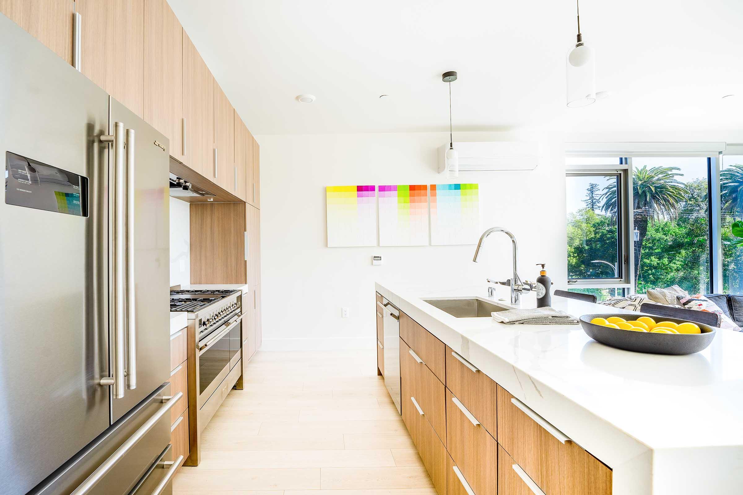 A modern kitchen featuring stainless steel appliances, wooden cabinetry, and a large sink. A colorful abstract painting hangs above the countertop, while a bowl of lemons sits on the counter. Large windows offer a view of palm trees and lush greenery outside, creating a bright and inviting atmosphere.