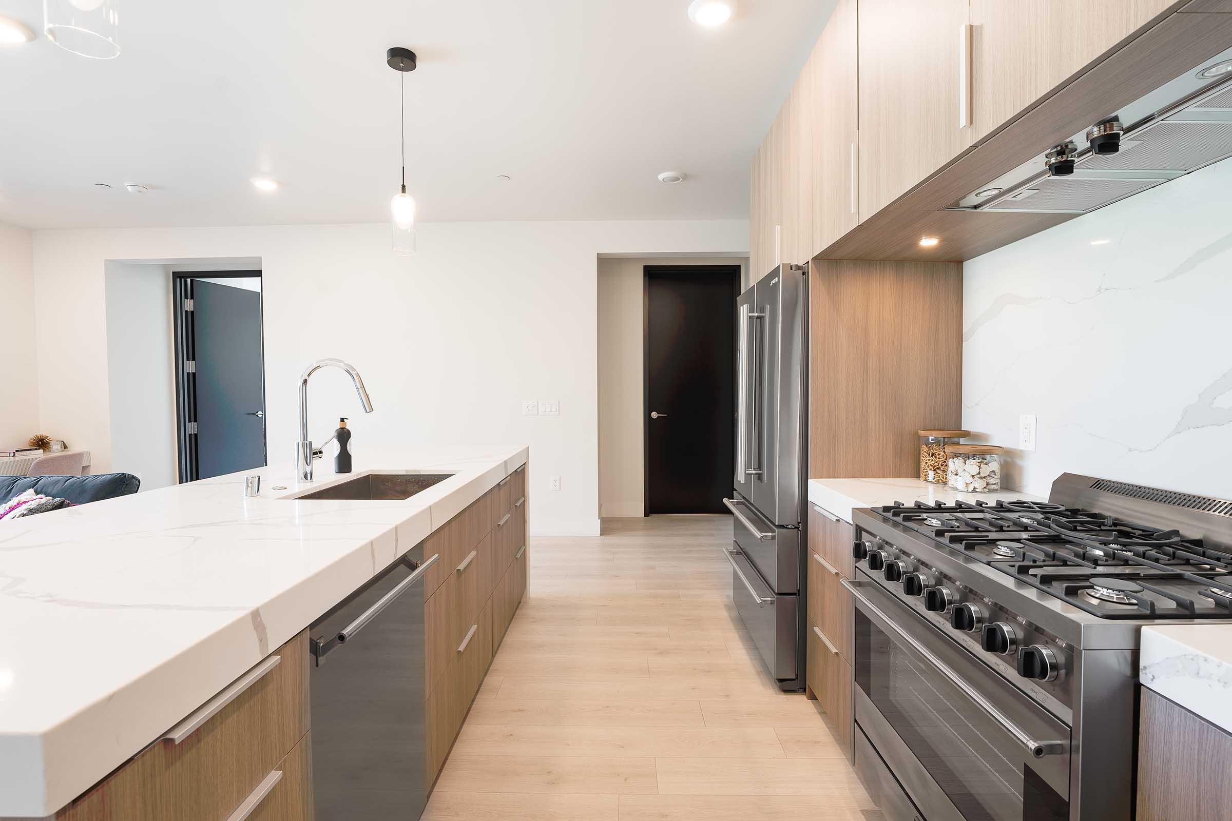 Modern kitchen with a large island featuring a white countertop, wooden cabinetry, and stainless steel appliances. A gas stove is visible on the right, while the left side shows a sleek faucet. The space has a bright, open design with light-colored walls and flooring, complemented by soft lighting fixtures.
