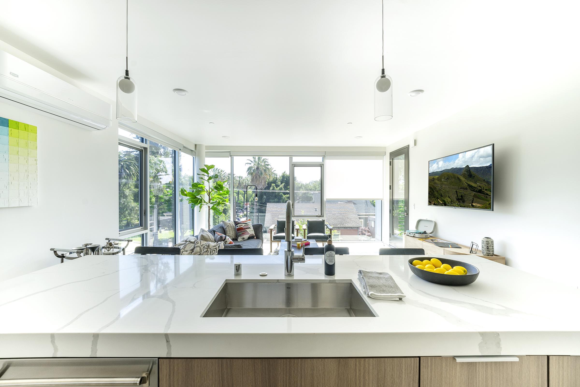 A modern kitchen featuring a marble countertop with a sink, a bowl of lemons, and stylish cabinetry. Large windows provide natural light and showcase a view of a living area with plants and comfortable seating. It includes sleek light fixtures and a wall-mounted TV.