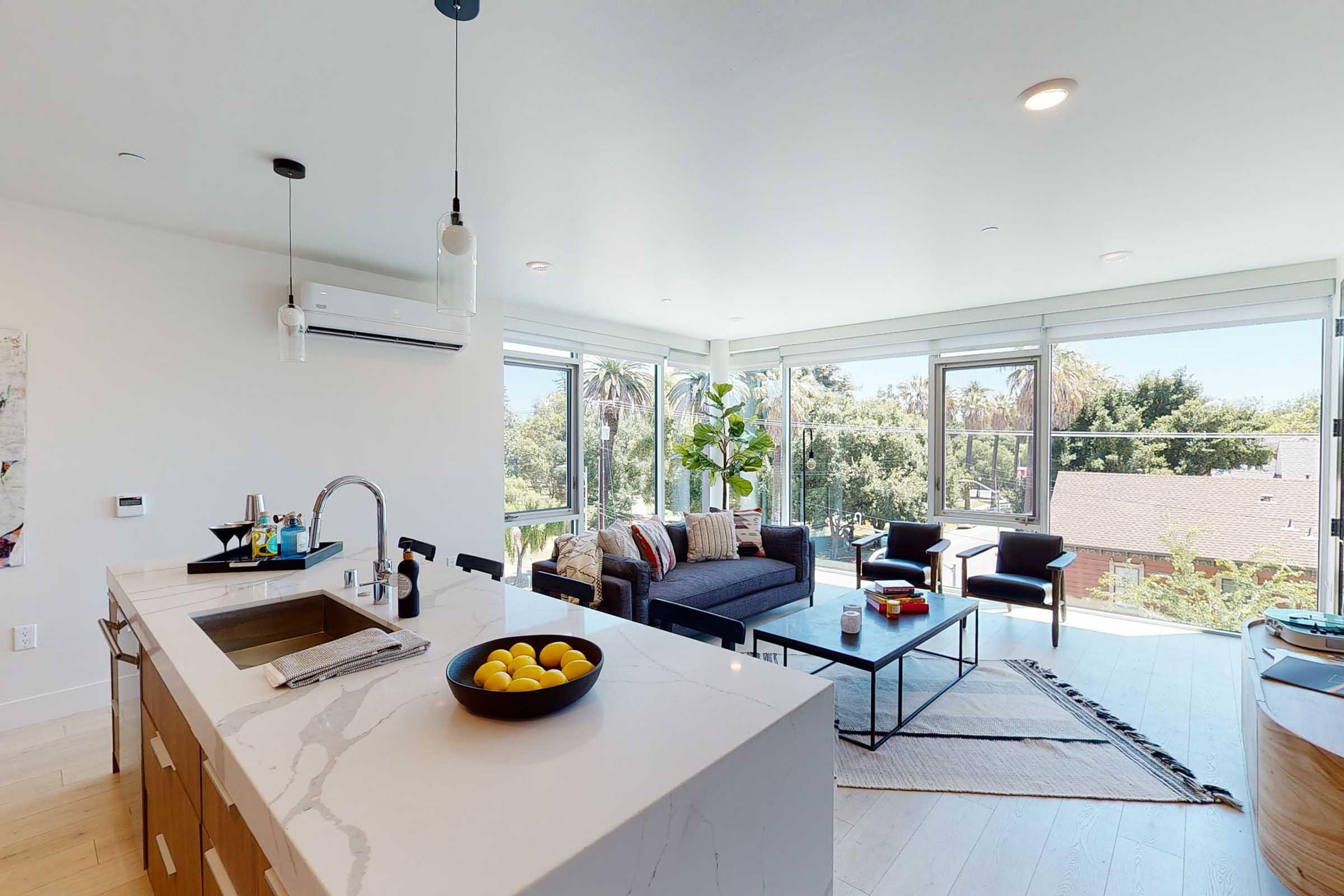 Bright and modern living space featuring a kitchen island with a sink and bowl of lemons, a cozy seating area with a sofa and coffee table, and large windows offering natural light and views of greenery outside. Decor includes a rug and potted plants, creating a welcoming atmosphere.