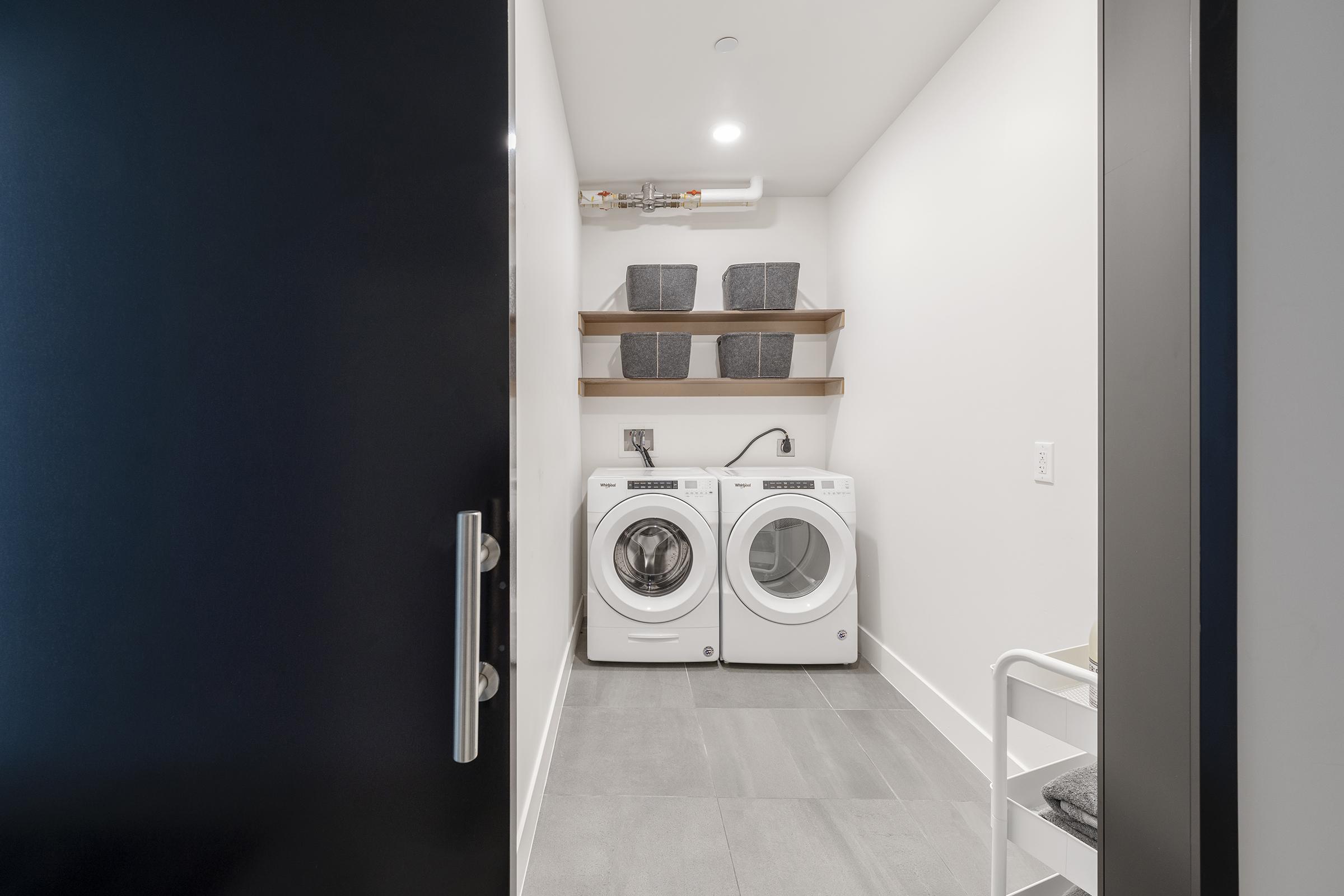 A modern laundry room viewed through an open black door, featuring a white washer and dryer stacked side by side. Above them, there are two wooden shelves holding gray storage bins. The space is well-lit with tiled flooring and a minimalist design.
