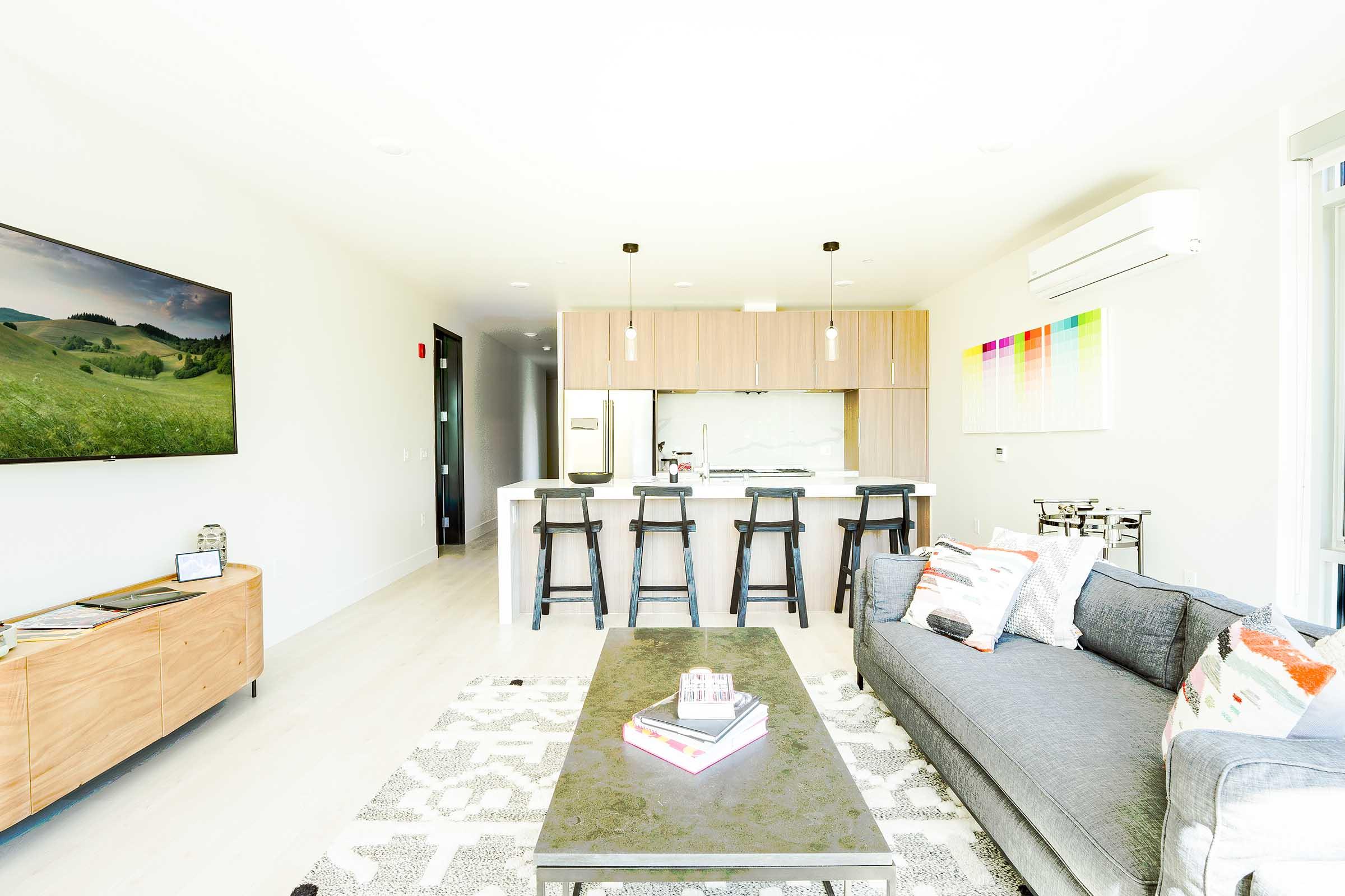 A modern living room with a grey sofa and a black coffee table. There is a large TV mounted on the wall and a sleek kitchen area in the background, featuring wooden cabinets and bar stools. Bright natural light fills the space, and colorful artwork decorates the wall. A cozy atmosphere with minimalistic decor.