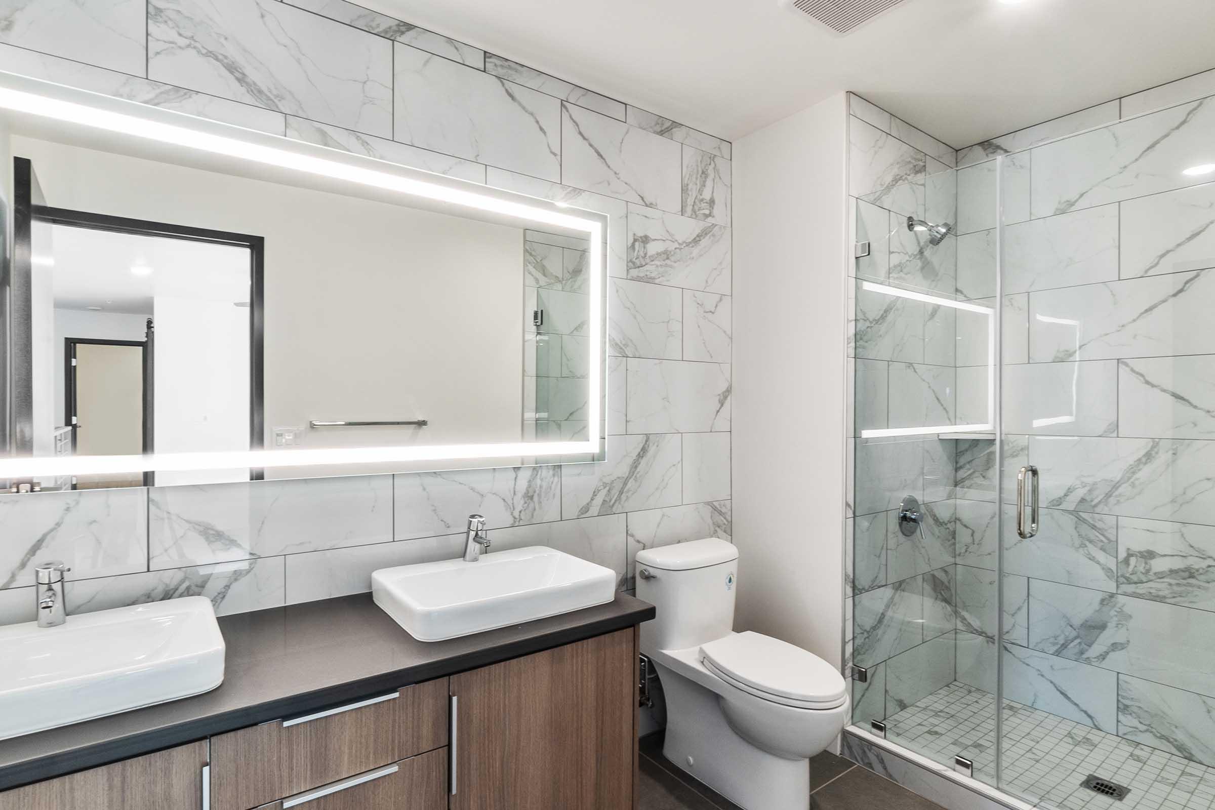 Modern bathroom featuring a large mirror with backlighting, two undermount sinks on a dark countertop, a white toilet, and a glass-enclosed shower with marble-patterned tiles. The sleek design is complemented by neutral tones and an overall spacious layout.