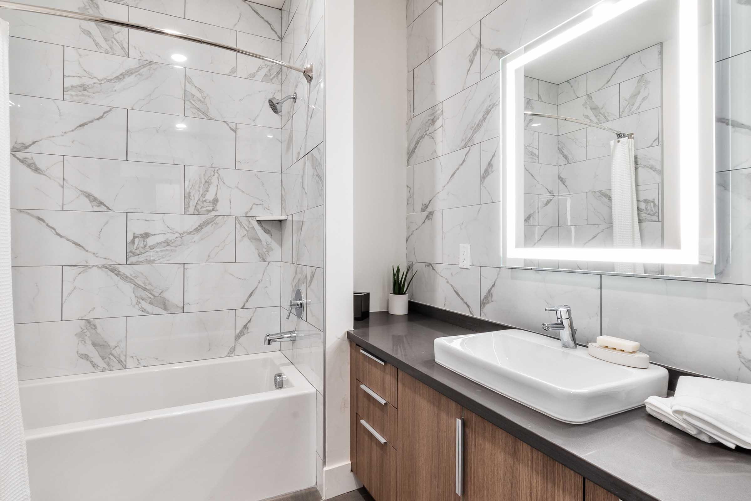 A modern bathroom featuring a bathtub with a shower, elegant marble tile walls, a sleek vanity with a large illuminated mirror, a white sink, and minimalist decor. The color palette combines neutral tones with subtle green accents from small plants.