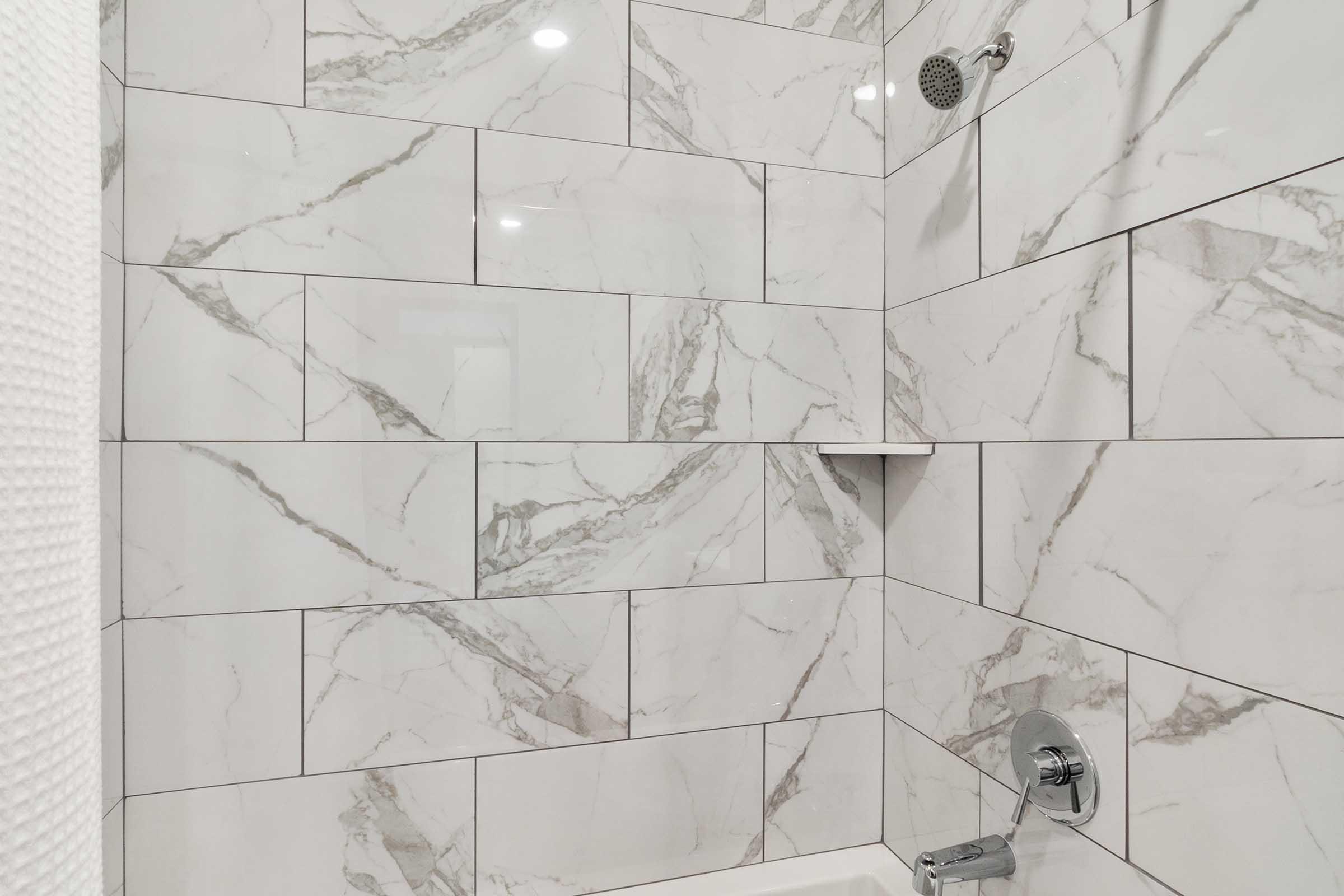 A modern shower space featuring sleek white marble tiles with gray veining. The walls are lined with large rectangular tiles, and there is a wall-mounted showerhead. A small shelf is built into the wall for storage. The overall design exudes a clean and contemporary aesthetic.