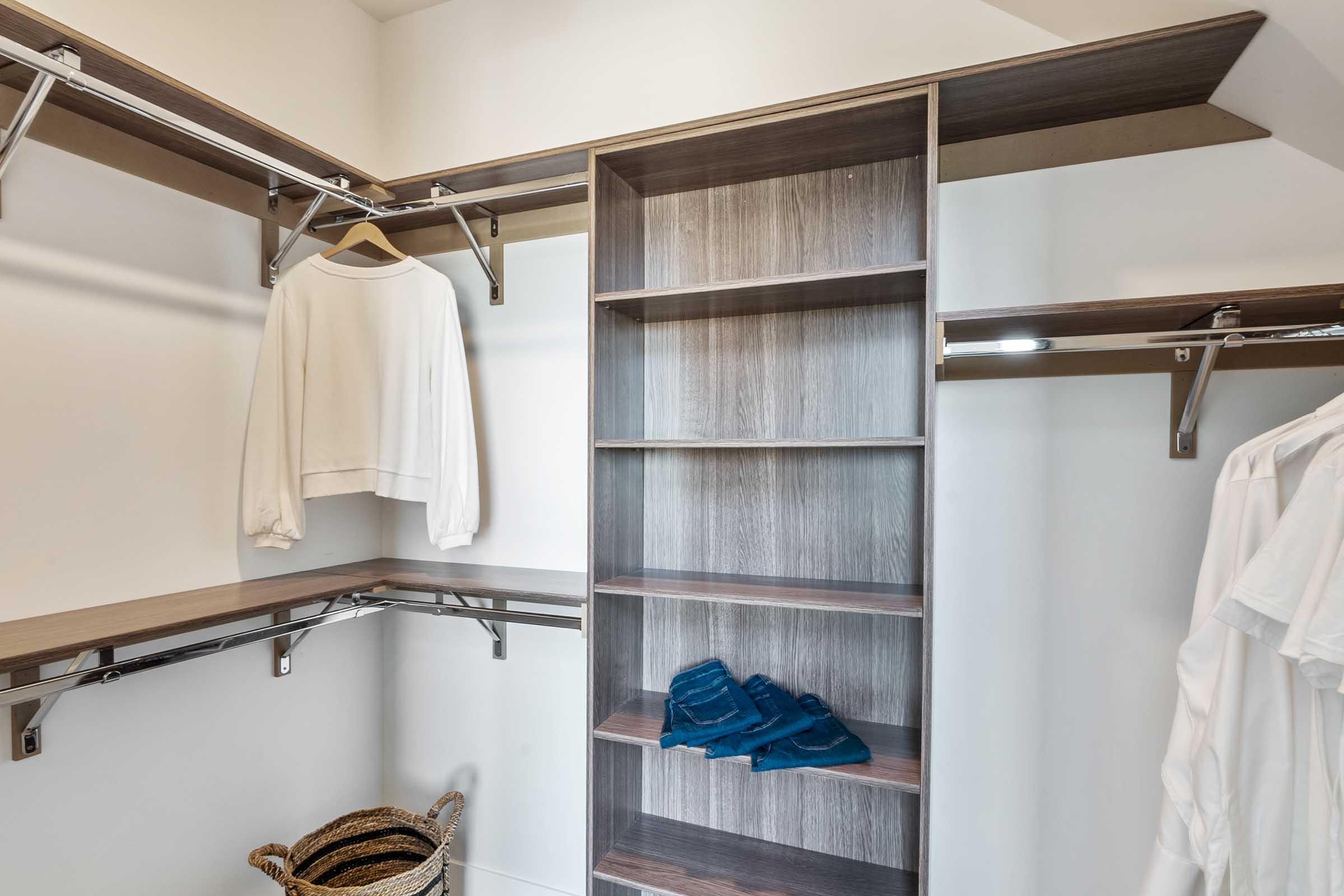 A minimalist closet featuring shelving and hanging space. There is a white long-sleeve shirt hanging on the left, with several stacked blue towels on the lower shelf. A woven basket sits on the floor, and the walls are clean and light-colored, creating an organized and airy feel.