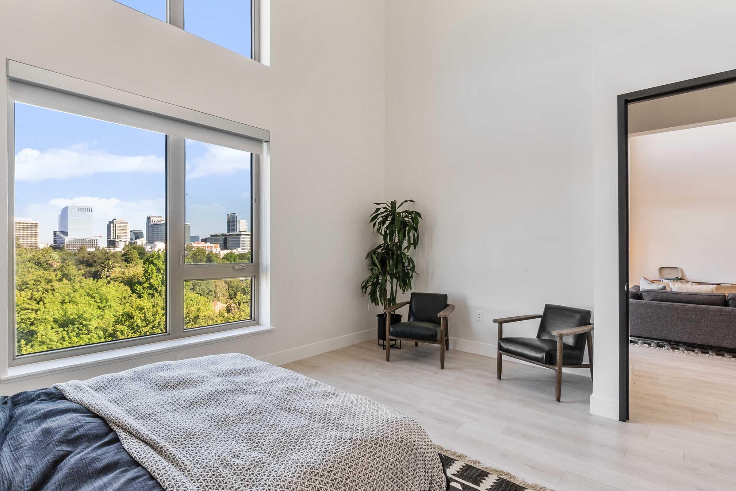 A modern bedroom featuring a large window displaying a city skyline and greenery outside. The room includes a bed with a textured blanket, two armchairs, and a potted plant, creating a bright and airy atmosphere. A doorway visible leads to a living area with a sofa.