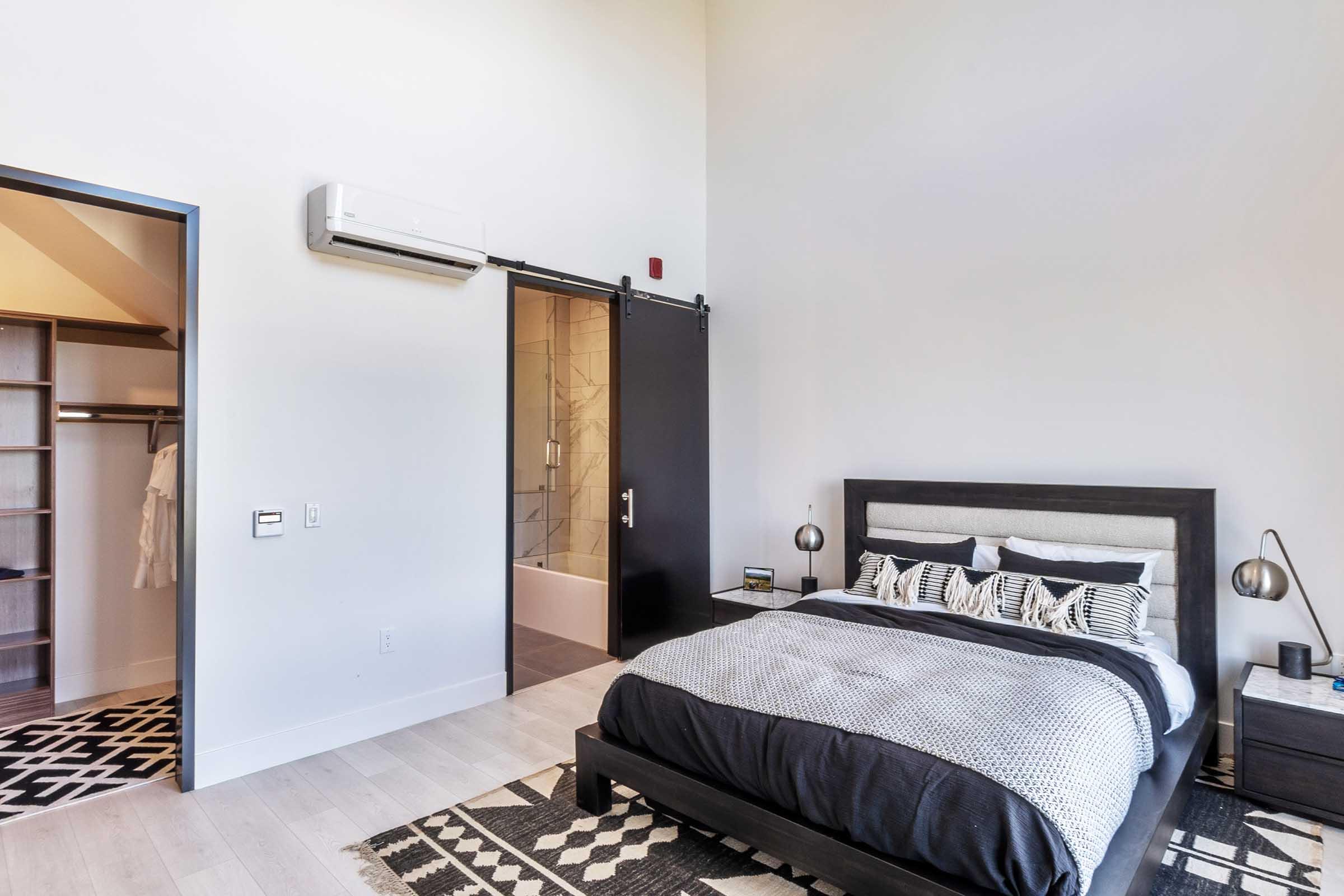 A modern bedroom featuring a large black framed bed with patterned bedding, a geometric rug, and stylish bedside lamps. The room has high ceilings, light walls, and a minimalist design, with a sliding door leading to a bathroom and a closet area visible in the background.