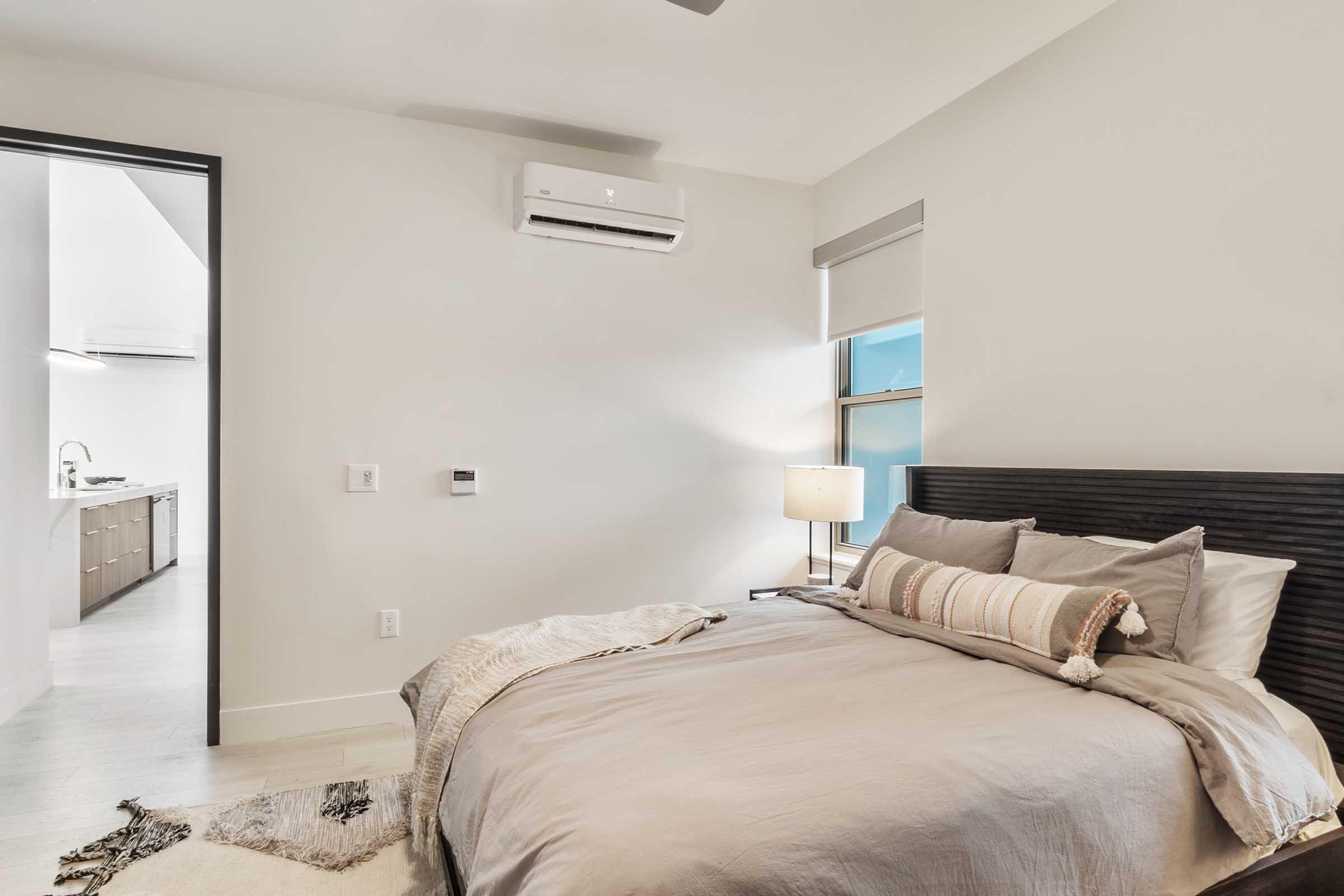 A cozy bedroom featuring a large bed with neutral-toned bedding, a bedside lamp, and a textured throw blanket. The walls are light-colored, and there is a window with soft natural light. A doorway in the background leads to a modern kitchen area.