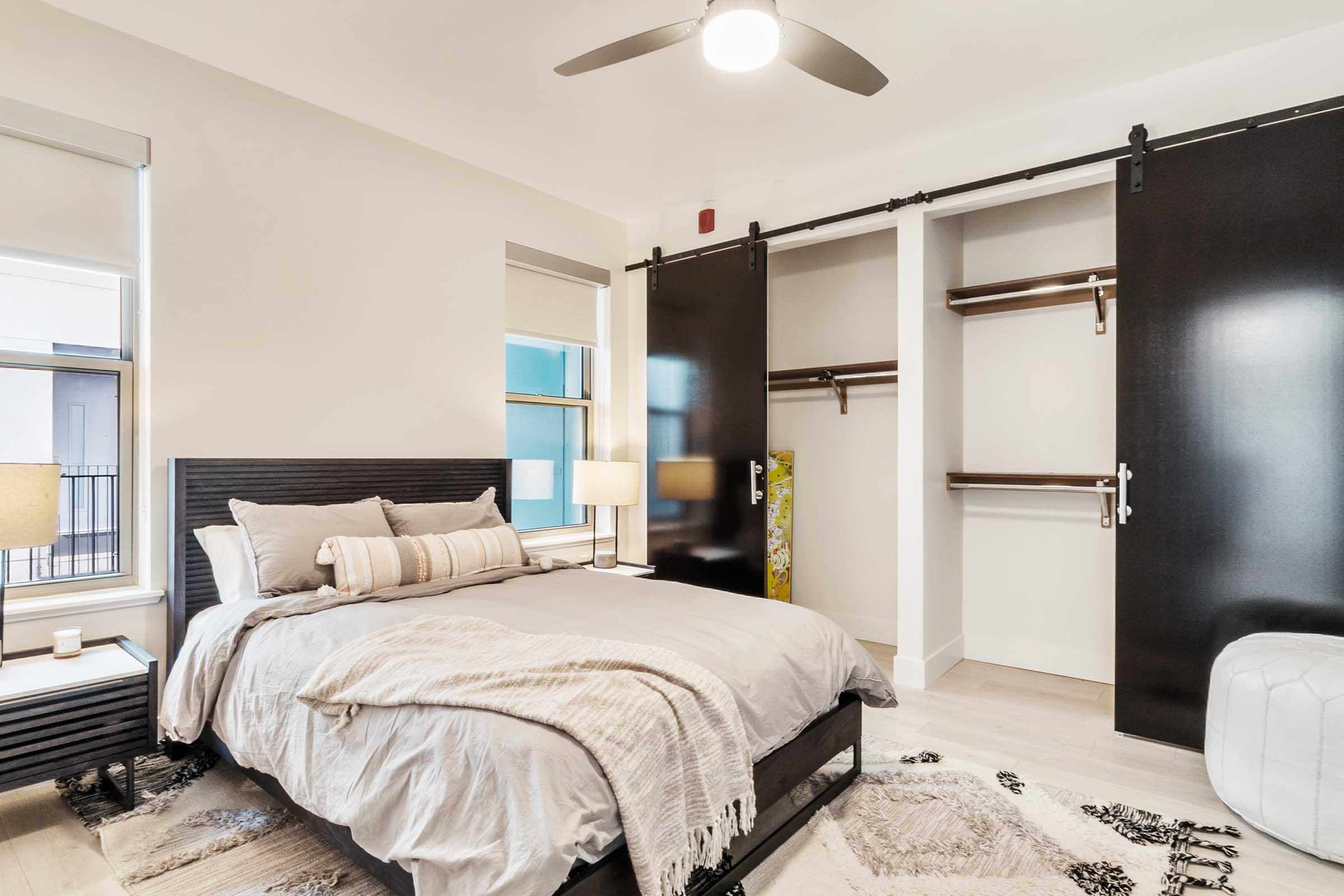 A modern bedroom featuring a queen-sized bed with light gray bedding and decorative pillows. There are two lamps on nightstands, a white ottoman at the foot of the bed, and a sliding black barn door revealing an empty closet space. The room has large windows allowing natural light in, with a neutral color palette throughout.