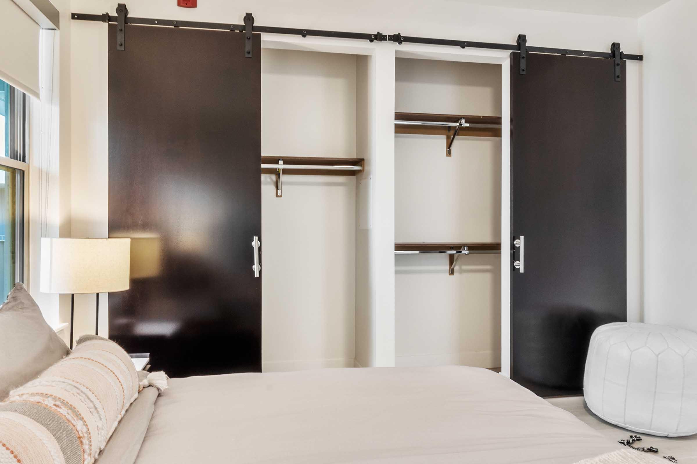 A modern bedroom featuring a neatly made bed with a cozy throw pillow. To the side, there are two black sliding closet doors open to reveal empty shelves and hanging space. A stylish lamp beside the bed and a white pouf enhance the contemporary decor. Natural light streams in through a nearby window.
