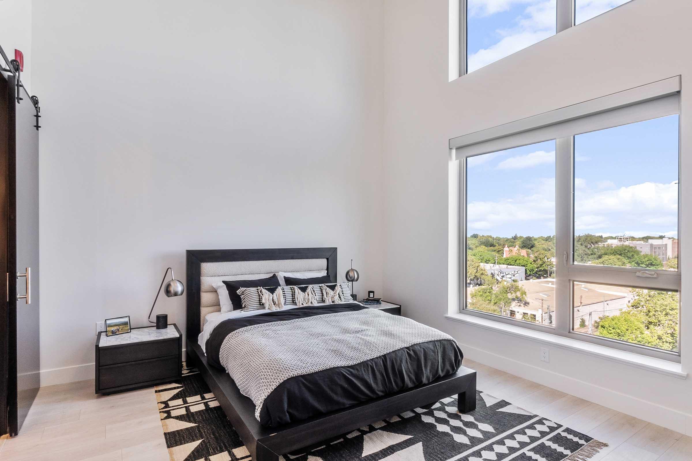 A modern bedroom featuring a black and white color scheme. The room has a large bed with decorative pillows, a nightstand with a lamp, and a patterned rug. Large windows allow plenty of natural light, showcasing a view of trees and buildings outside. The walls are painted in a light color, enhancing the spacious feel.