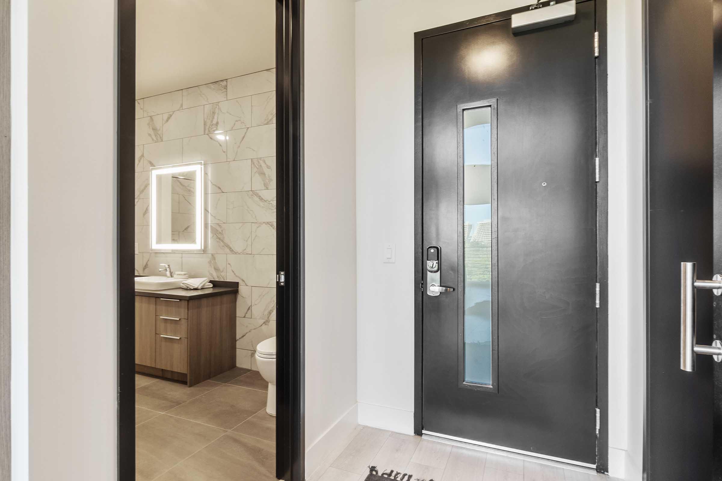 A modern entryway featuring a black door with a glass panel, leading to a bathroom with marble walls and a stylish vanity. The space is well-lit, showcasing a clean and contemporary design, with a tiled floor and a welcome mat near the entrance.