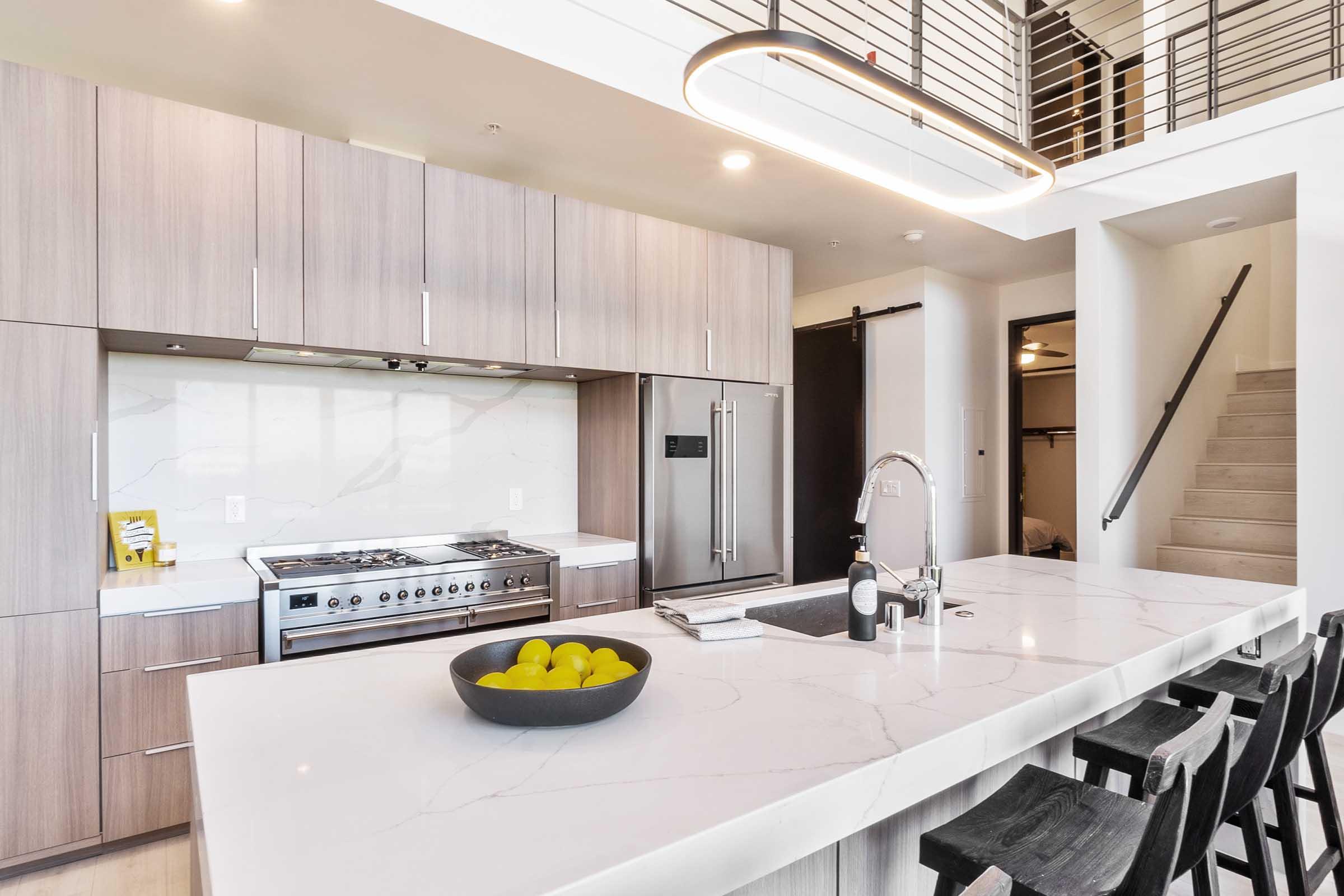 Modern kitchen featuring sleek cabinetry in warm wood tones, a large marble island with a bowl of green apples, stainless steel appliances including a gas stove, and an open layout with a spiral staircase in the background. Bright and airy atmosphere enhanced by natural light.