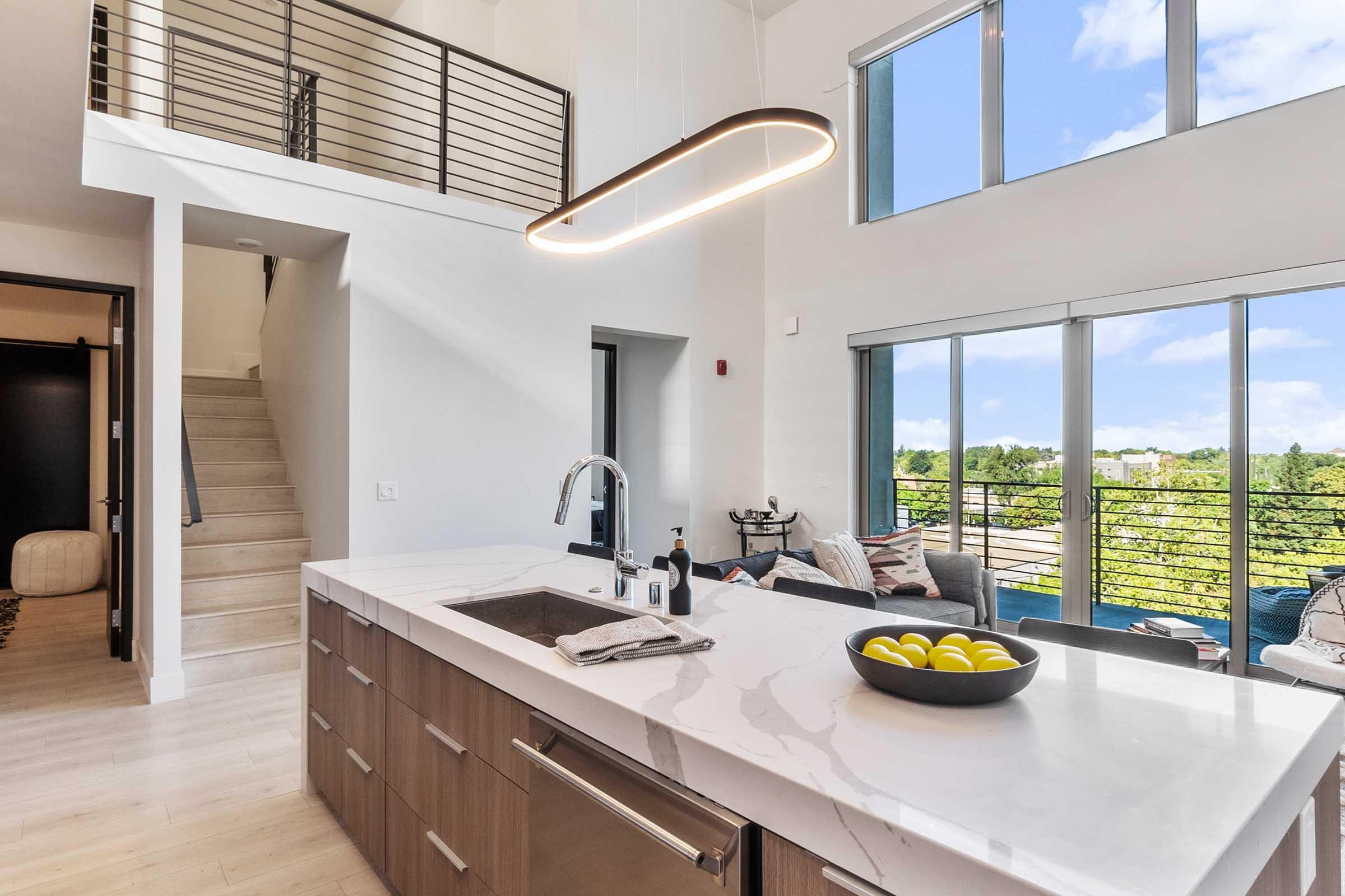 Modern kitchen featuring a large marble island with a bowl of yellow apples, sleek wooden cabinets, and a stylish pendant light. Large windows allow natural light to fill the space, showcasing a view of greenery outside. A staircase leads to an upper level, enhancing the open and airy atmosphere