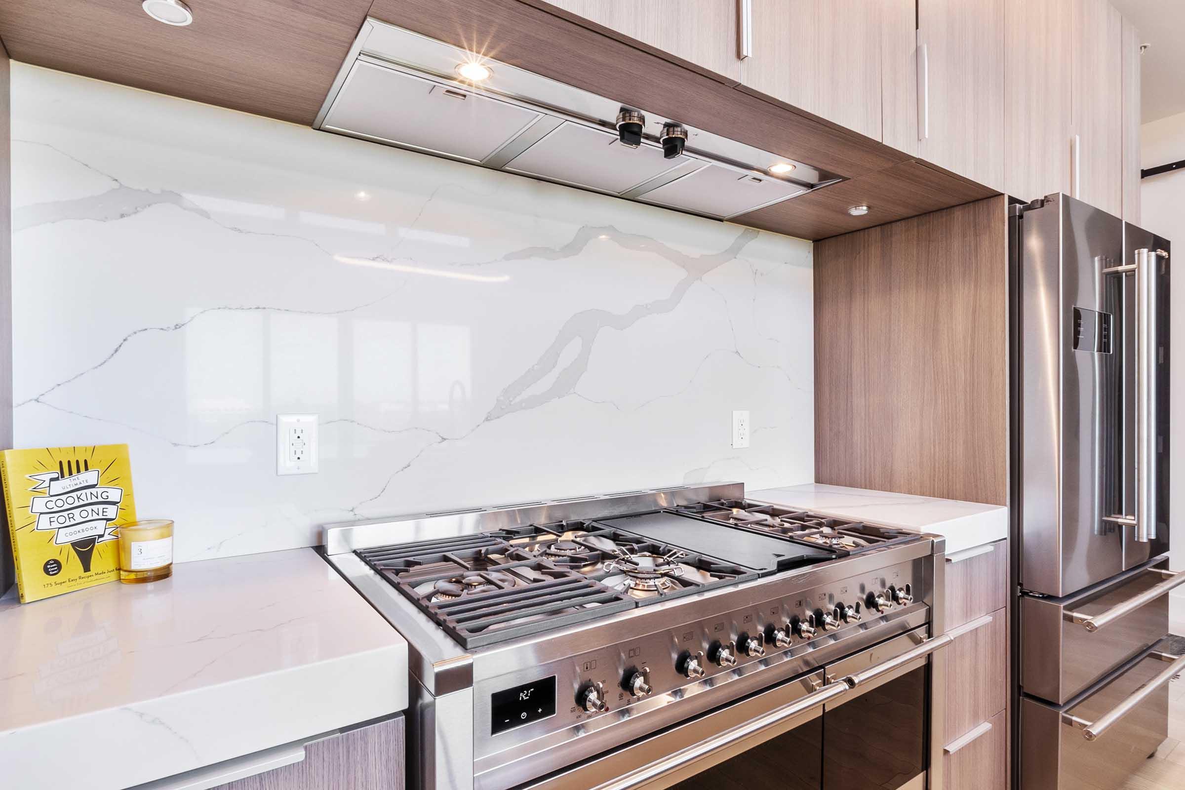 A modern kitchen featuring a stainless steel gas stove, sleek countertop, and a marble backsplash. On the counter, there's a yellow cookbook titled "Cooking for One" and a candle. The cabinetry is a mix of light and dark wood, and there's a stainless steel refrigerator visible.