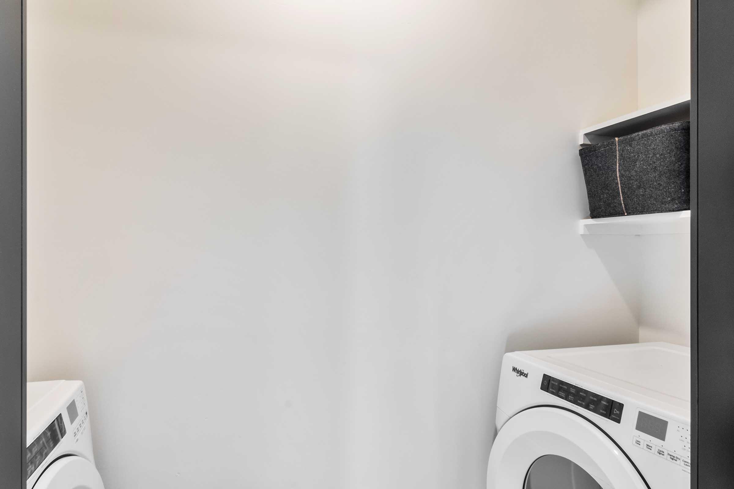 A small laundry room featuring a white washing machine and dryer side by side. Above the appliances, there is a shelf containing a dark storage basket. The walls are plain and the overall space appears bright and uncluttered.