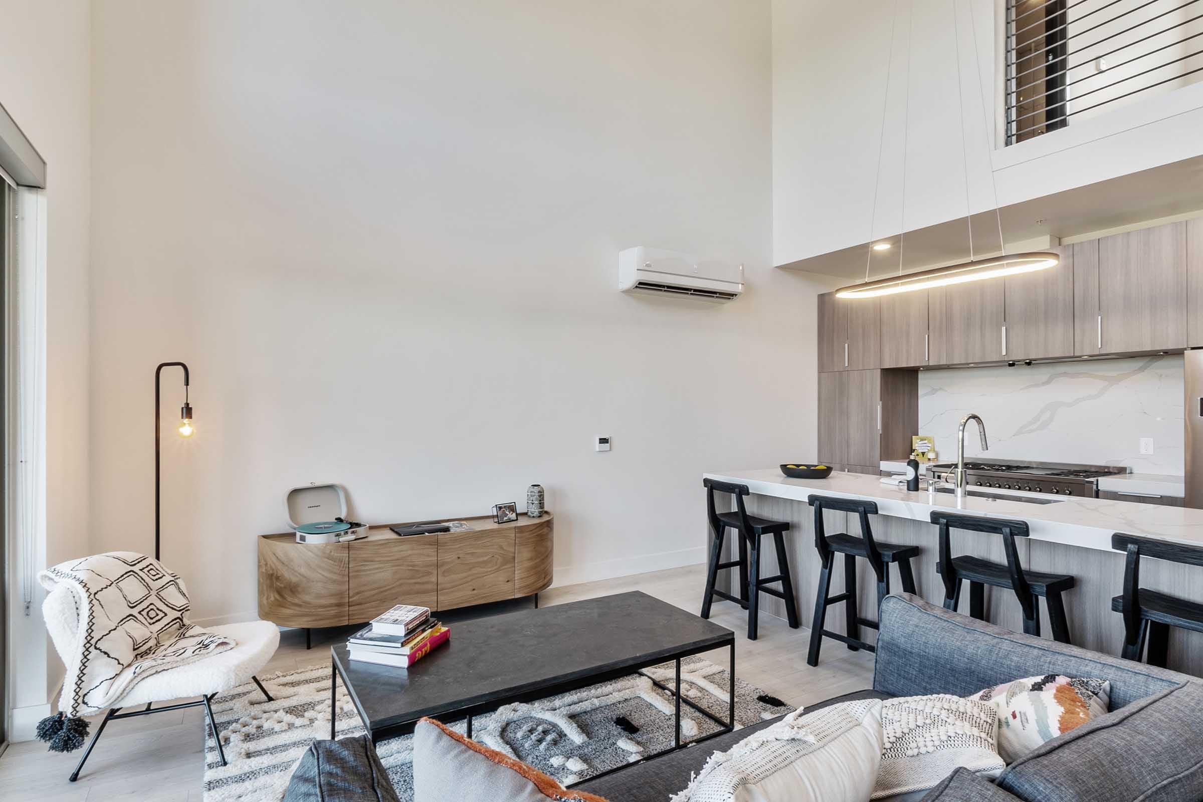 A modern, stylish living space featuring a cozy sofa, a black coffee table, and a wooden sideboard. The kitchen is visible in the background, with sleek cabinetry and bar stools. Large windows let in natural light, enhancing the open, airy feel of the room.