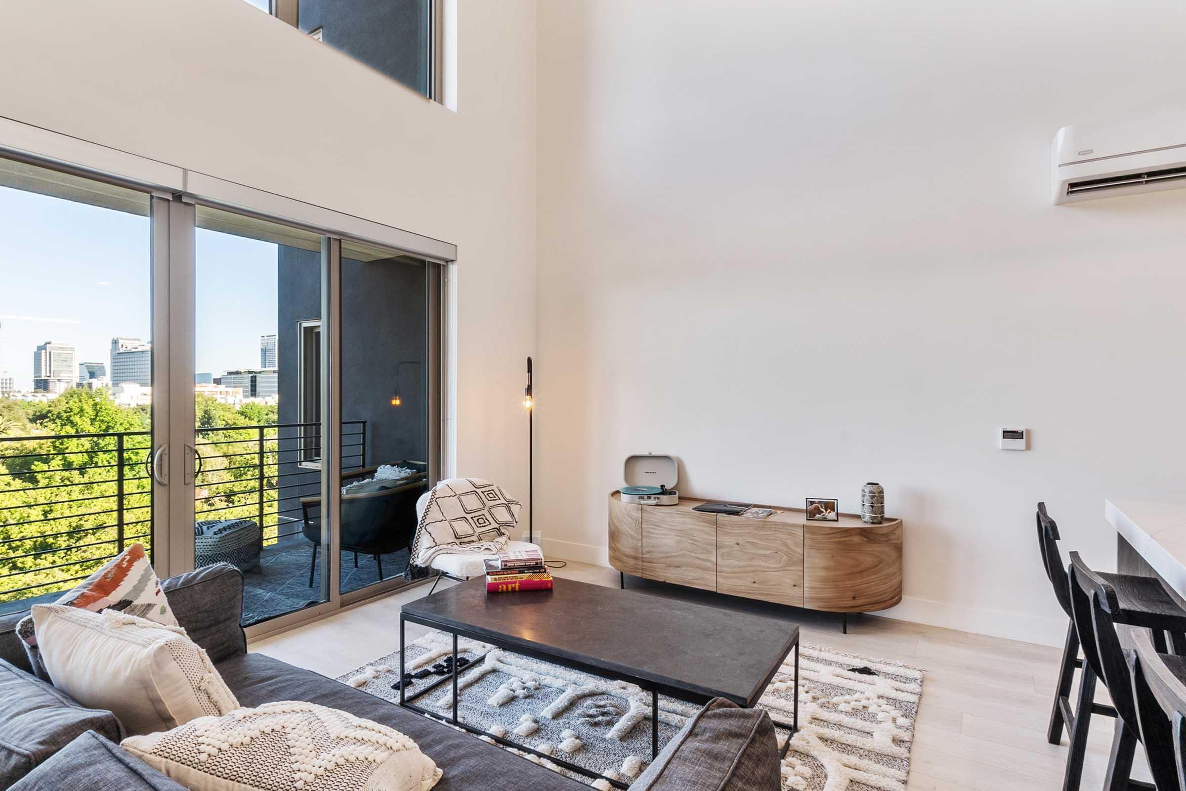 Modern living room with high ceilings, featuring a gray sofa, a black coffee table, and a wooden console unit. Large glass doors lead to a balcony with greenery visible outside. A stylish armchair and decorative items add a cozy touch to the space. Bright and airy atmosphere with natural light