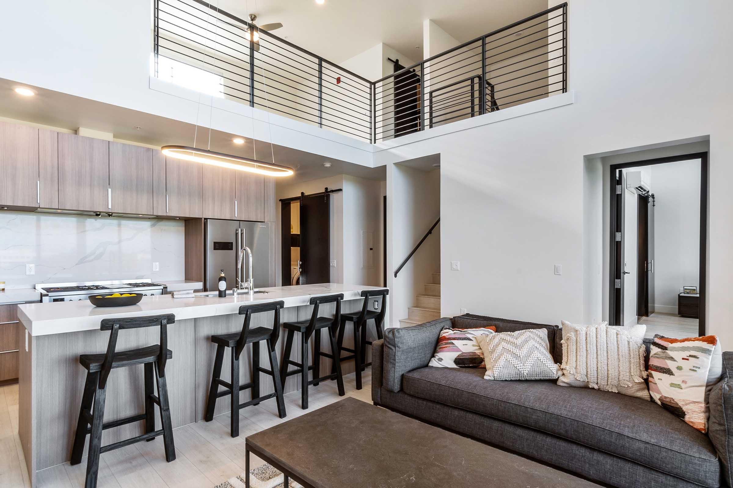 Modern open-concept living space featuring a kitchen with a central island and bar stools, adjacent to a cozy living area with a dark sofa adorned with decorative pillows. A sleek staircase leads to a lofted area above, all set within a bright, minimalist design.