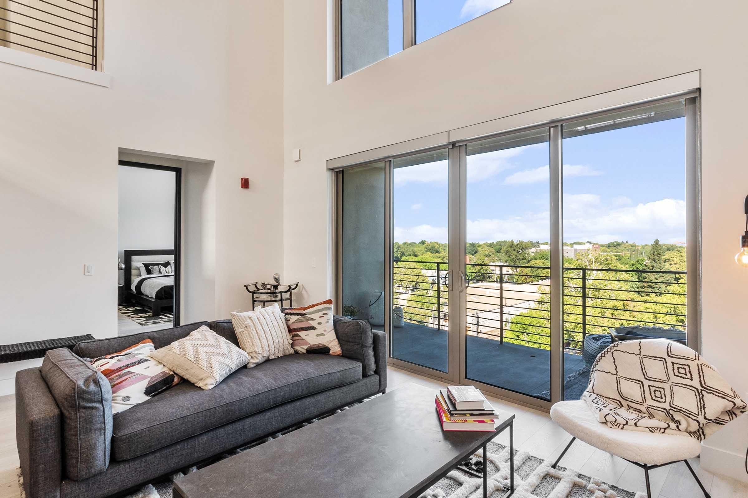 A modern living room featuring a dark gray sofa with patterned throw pillows, a coffee table with books, and a light-colored chair. Large windows offer a view of greenery outside, and there is a glimpse of another room in the background. The space is bright and airy with minimalist decor.