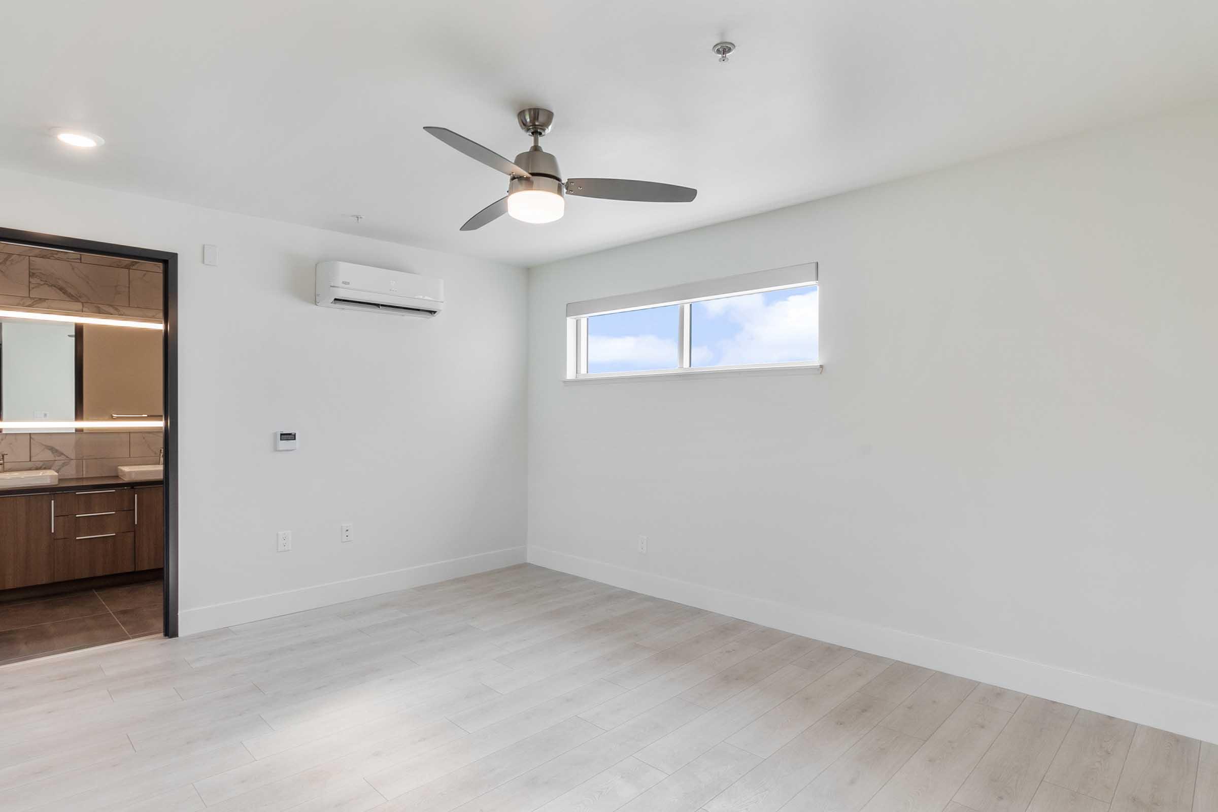 A bright, empty room with light-colored walls and flooring. It features a ceiling fan and a window allowing natural light. To the left, a doorway leads to another space, possibly a bathroom, indicated by wall-mounted fixtures and mirrors. The overall atmosphere is modern and minimalistic.