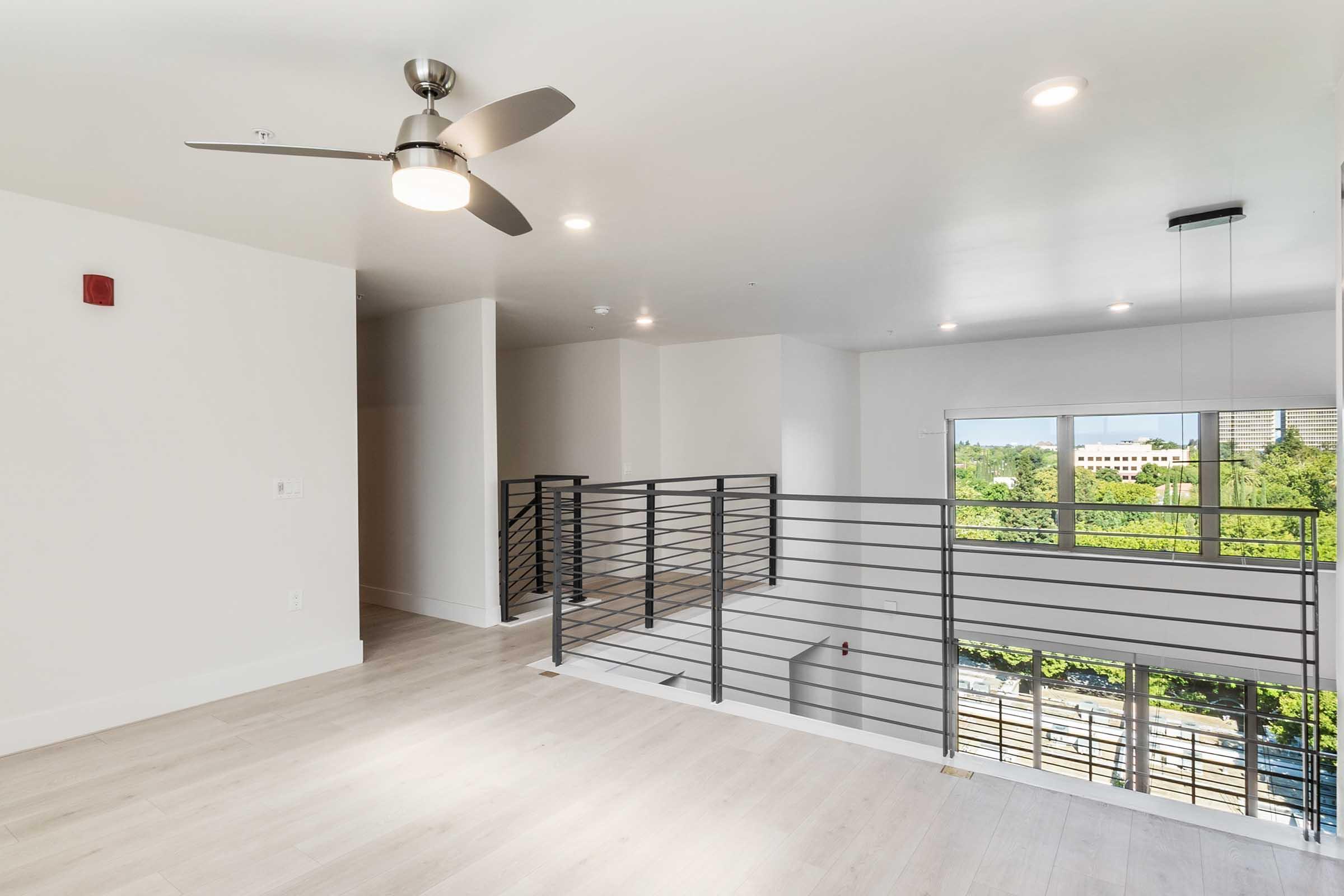 Spacious and modern interior of an apartment with light-colored wooden flooring, a ceiling fan, and large windows providing natural light. A contemporary railing separates the living area from a balcony view of greenery outside. The walls are painted in a light shade, creating a bright and airy atmosphere.