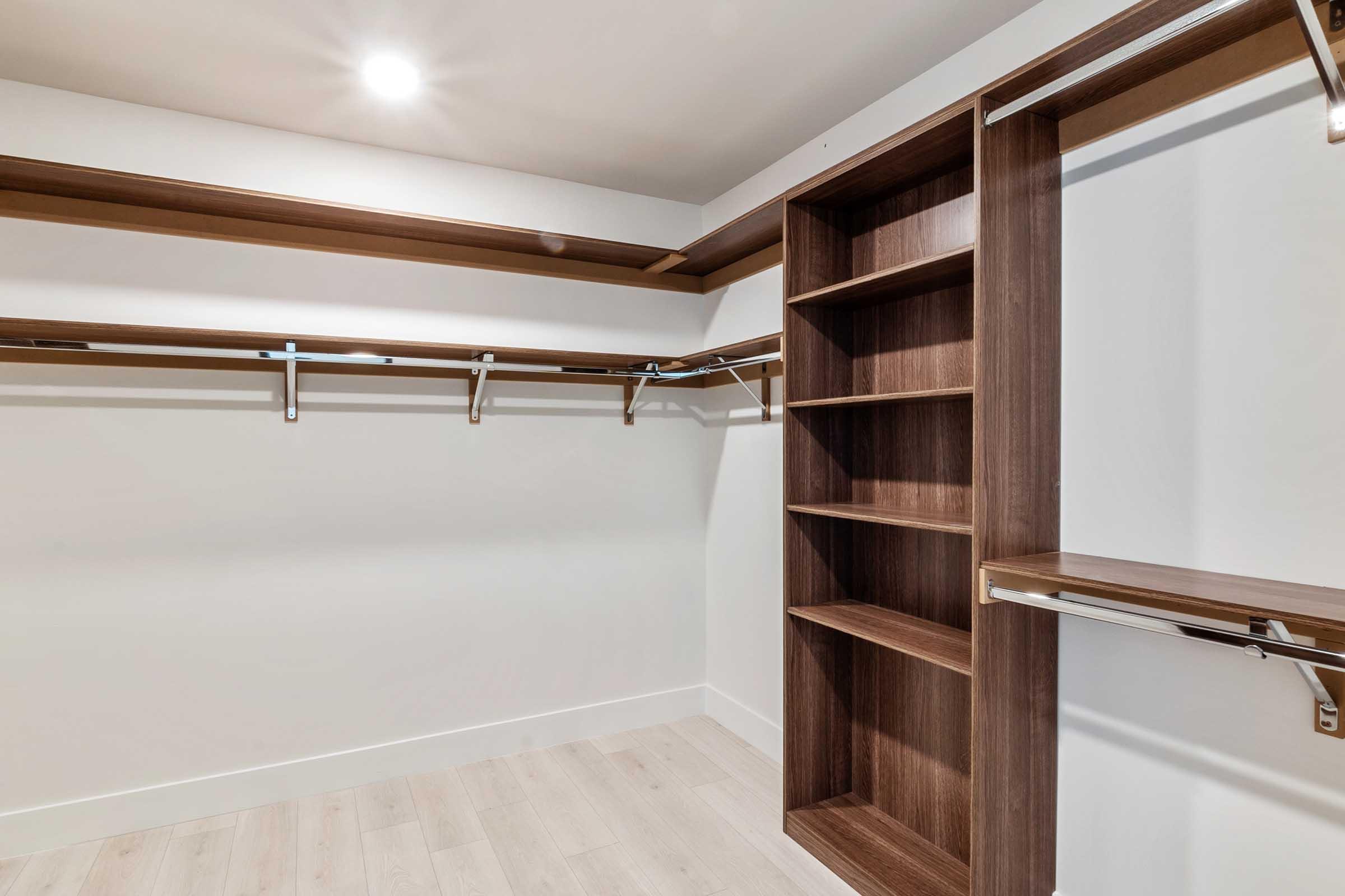 A spacious, empty closet with light-colored wood flooring. It features wooden shelves along the top, hanging rods on both sides, and a tall wooden shelving unit on the right, designed for organized storage. The walls are painted in a light shade, and there is ample natural light from a ceiling fixture.