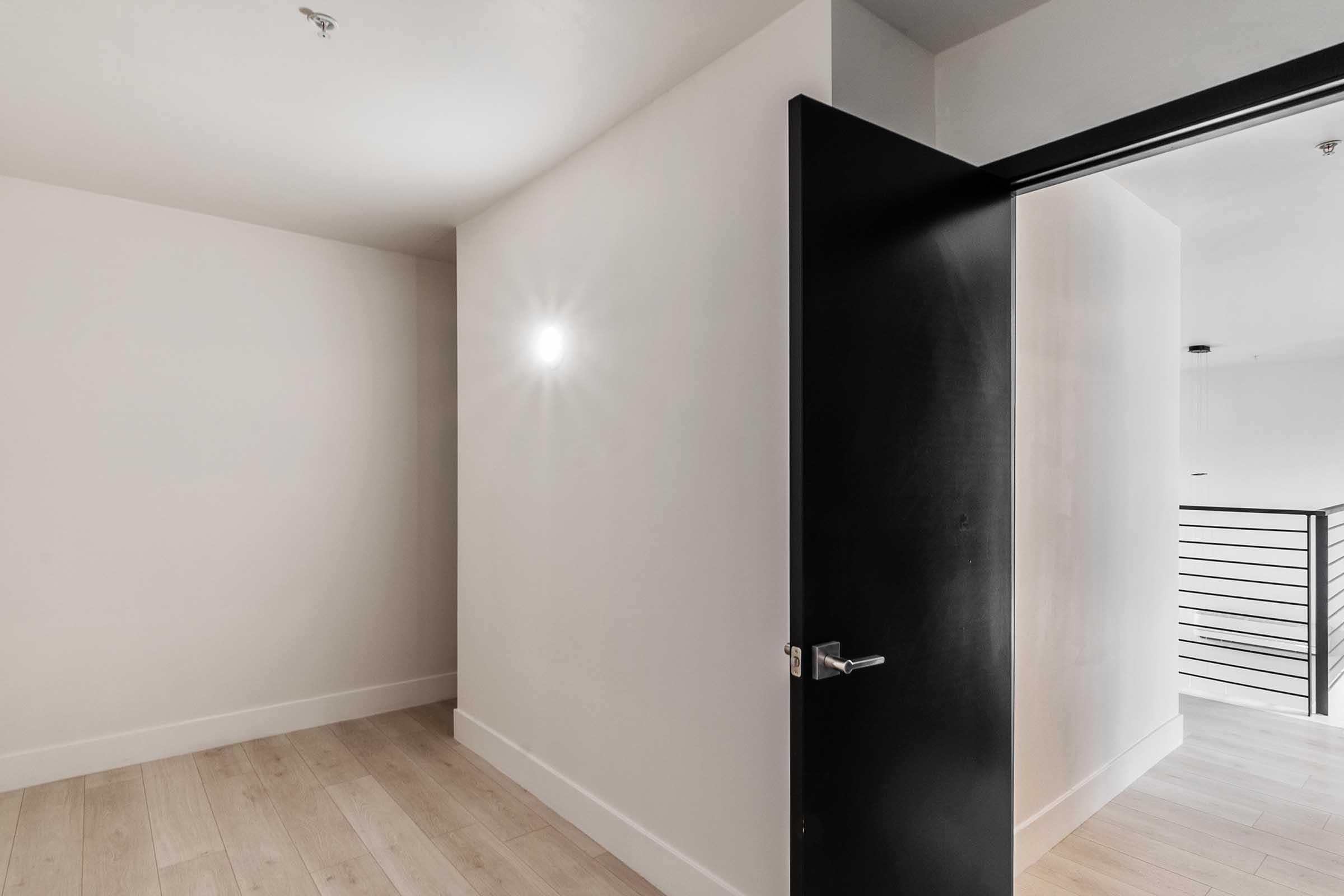 A minimalist interior space with light-colored walls and wooden flooring. A black door is partially open, leading to another area, and a small light fixture on the wall provides soft illumination. The overall atmosphere is modern and uncluttered.