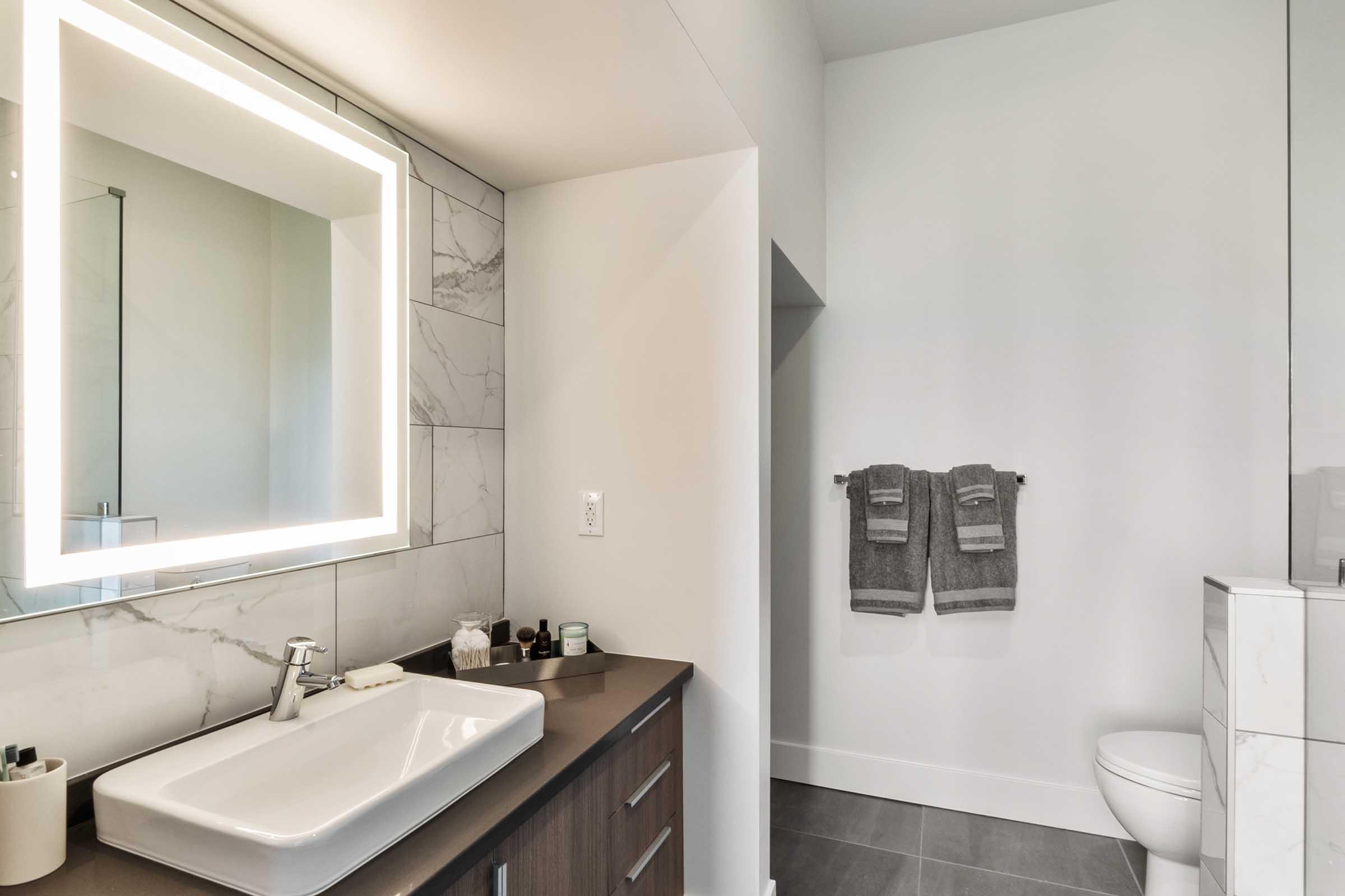 Modern bathroom with a sleek design featuring a large illuminated mirror above a white sink. There are brown cabinetry and neatly arranged toiletries on the counter, while gray towels hang on a rack nearby. A glass shower enclosure is visible, and the floor is tiled in gray.