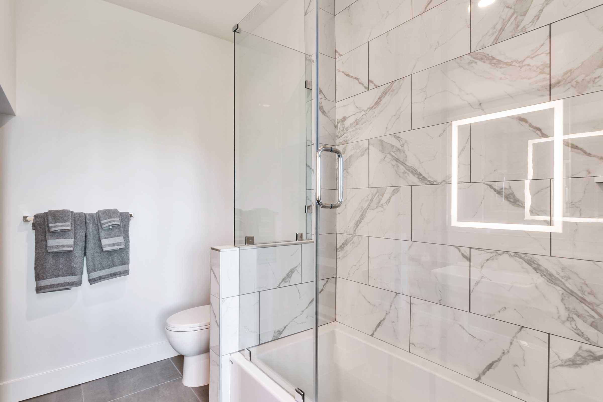 A modern bathroom featuring a shower with a glass enclosure, a bathtub, and marble tile walls. There are two gray towels hanging on a rack and a sleek toilet in the corner. Soft, ambient lighting is integrated into the design, enhancing the contemporary feel of the space.