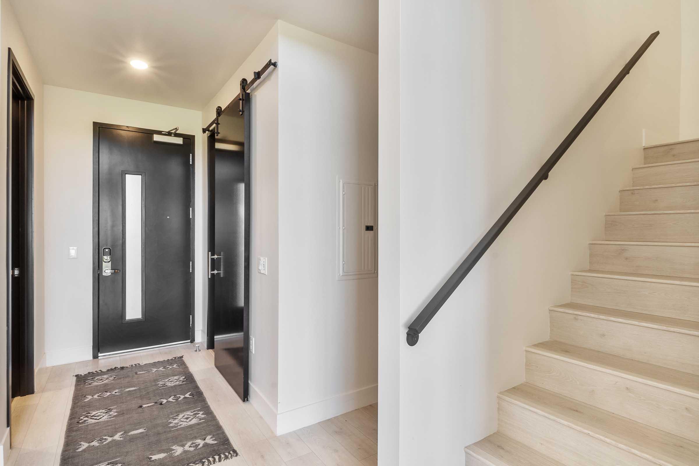 An entryway featuring a black front door with a glass panel, a dark mailbox mounted on the wall, a gray patterned runner rug on light wooden flooring, and a staircase with wooden steps and a metal railing leading to the upper level. Light streams through the space, creating a welcoming atmosphere.