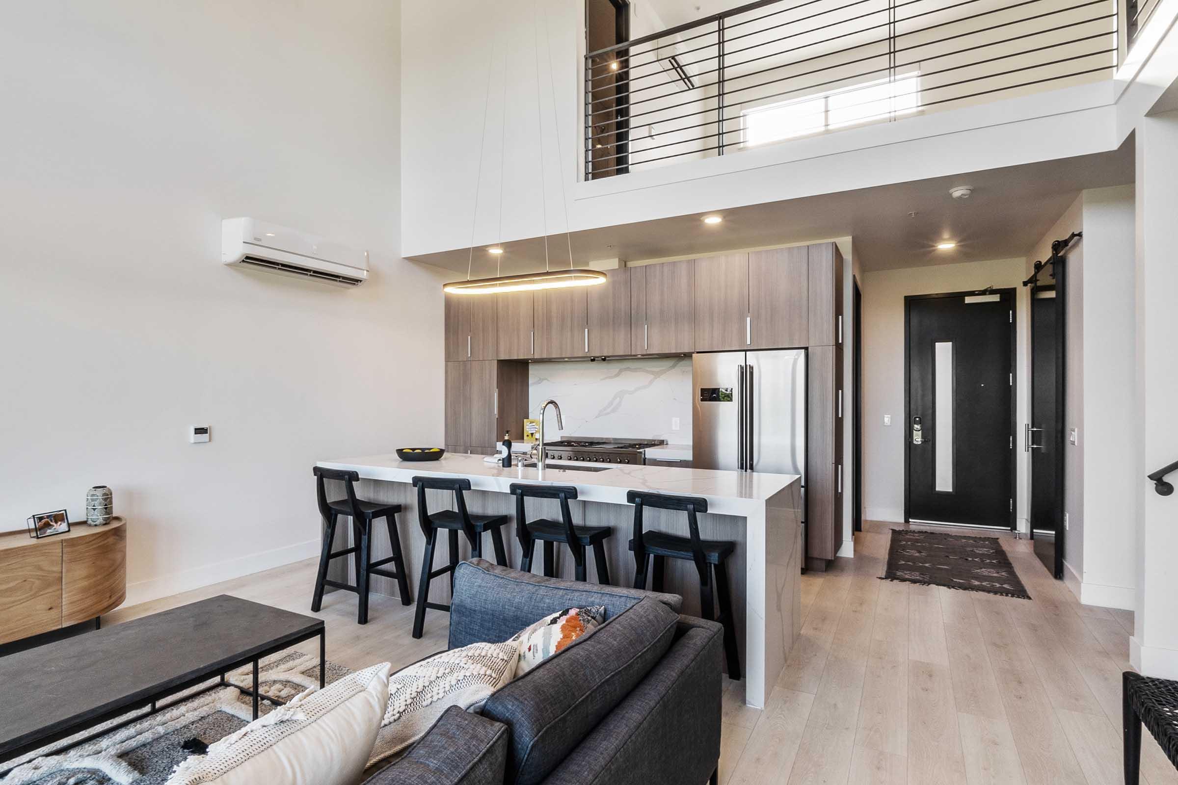 A modern open-concept kitchen and living area with a large island, barstool seating, and stainless steel appliances. The space features high ceilings, stylish decor, and a comfortable couch. There are large windows allowing natural light, and a staircase leading to a lofted area.