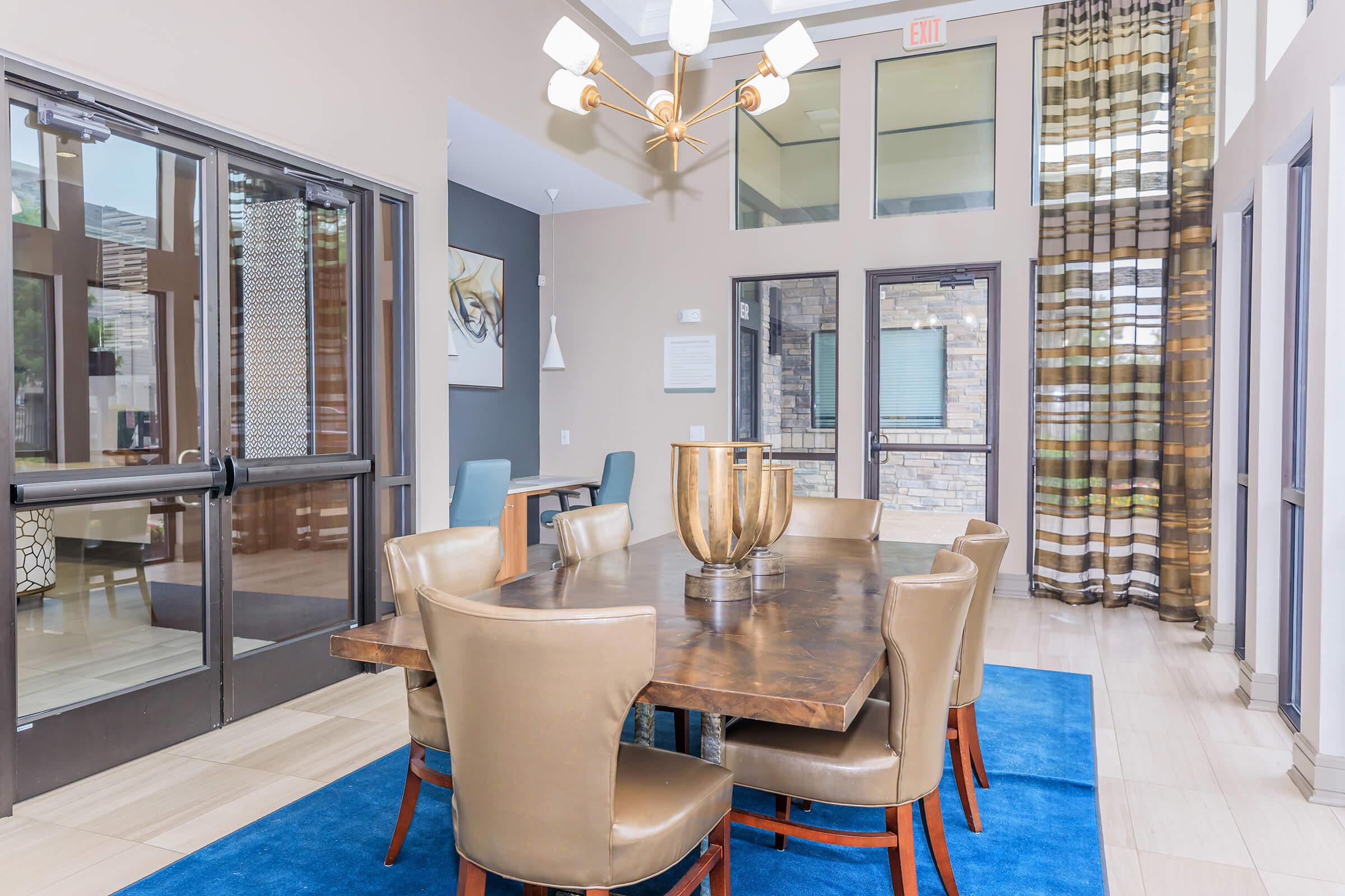 A modern dining area featuring a large wooden table surrounded by six tan chairs. A decorative bowl sits in the center of the table. Large windows with sheer and patterned curtains let in natural light, and a stylish chandelier hangs from the ceiling. Two doors lead to adjacent spaces.