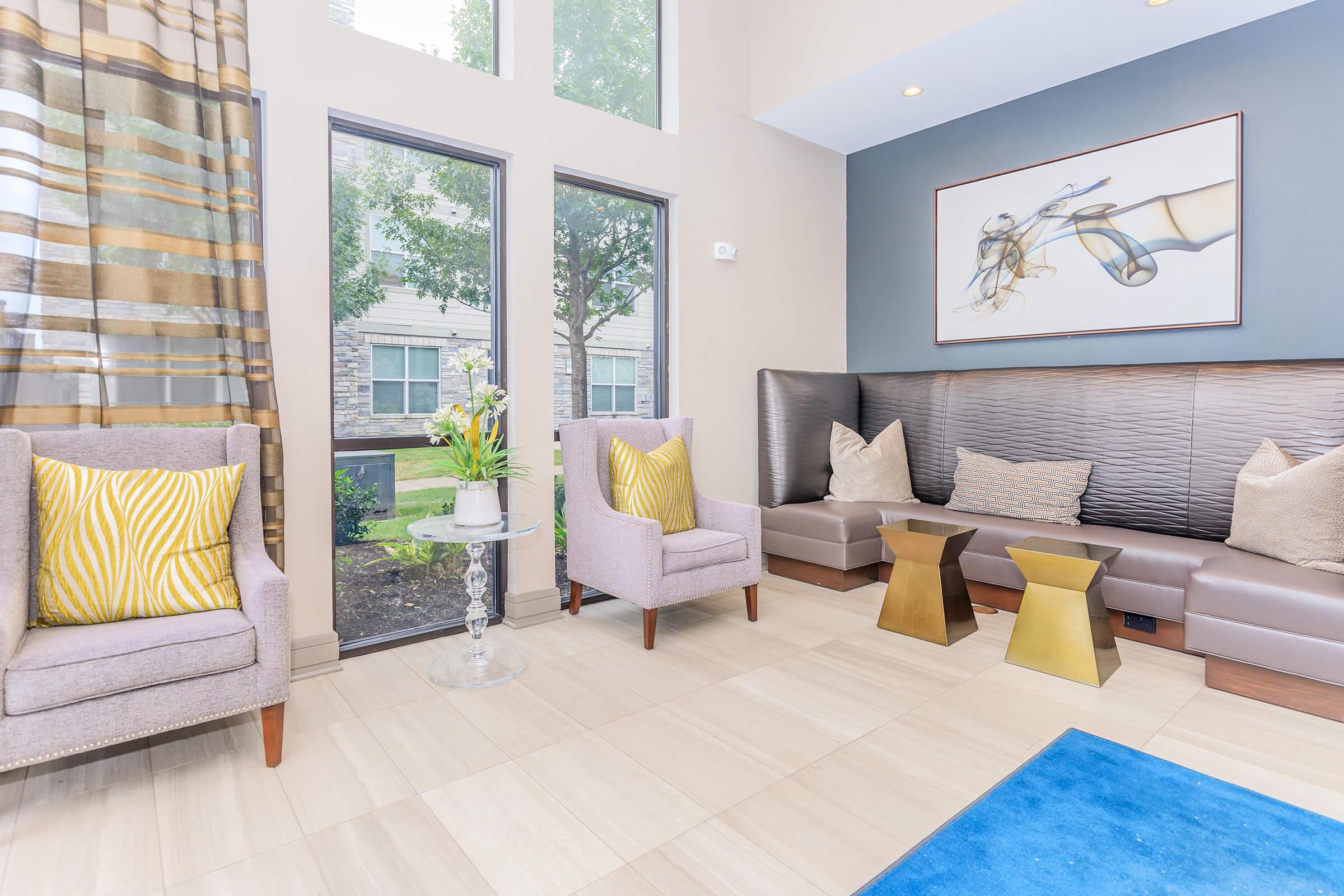 A modern lounge area featuring two light purple armchairs with yellow accent pillows, a small coffee table with gold bases, and a decorative plant. Large windows allow natural light to fill the space, complemented by neutral-colored walls and a piece of abstract art above the seating.
