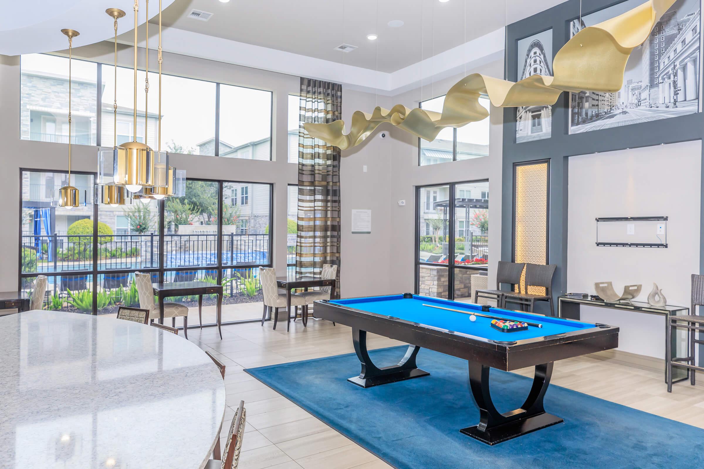Interior view of a modern lounge area featuring a blue billiard table, stylish light fixtures, and large windows allowing natural light. There are seating areas with elegant chairs and a decorative wall showcasing architectural photos. A swimming pool is visible outside through the windows.