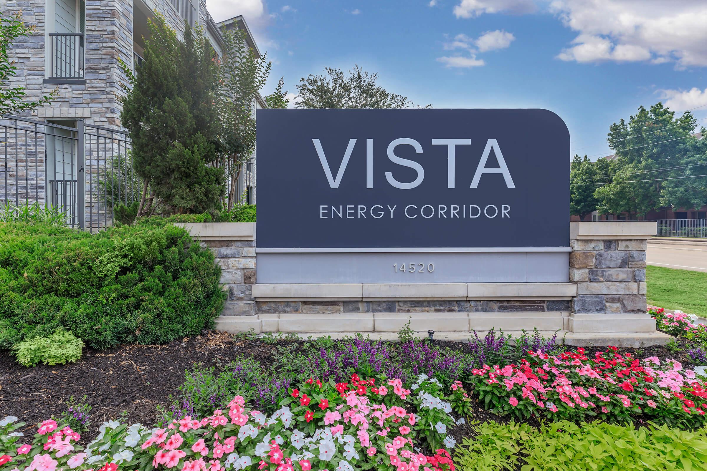 A sign for "VISTA ENERGY CORRIDOR" with the address 14520, surrounded by colorful flower beds and greenery, set against a stone background. The sky is partly cloudy, adding to the welcoming appearance of the location.