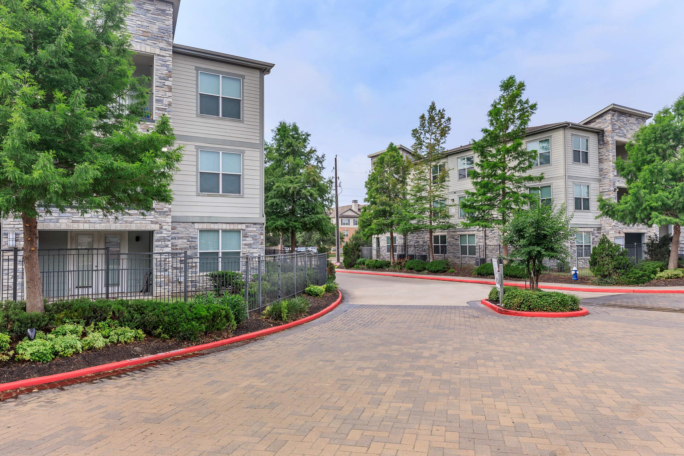 A landscaped residential area featuring two multi-story apartment buildings with stone accents. The pathway is paved with bricks, lined by neatly trimmed shrubs and trees, creating a welcoming entrance. The sky is slightly overcast, adding a serene atmosphere to the scene.