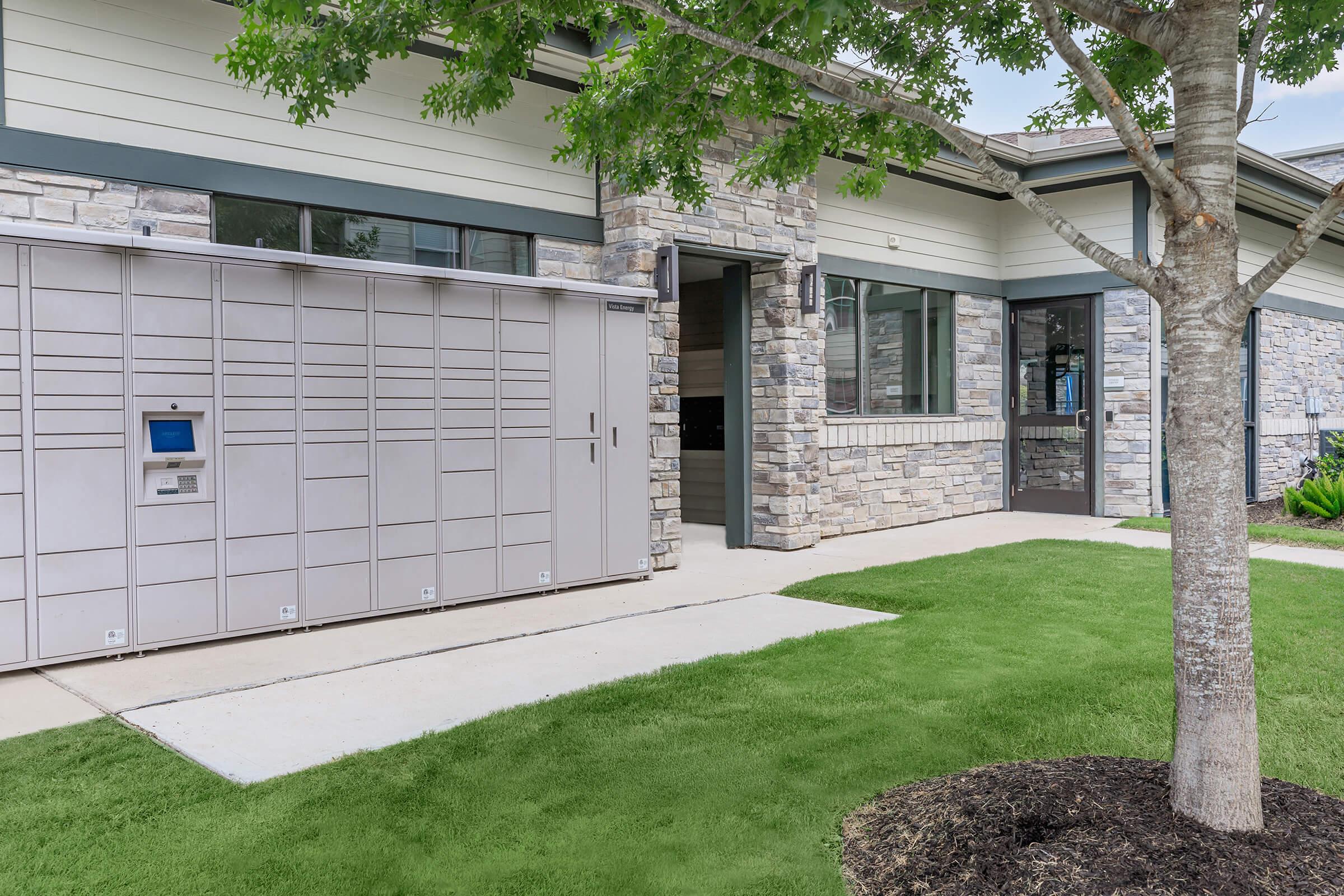 A modern building facade featuring a stone wall and a large parcel locker system near the entrance. The area is well-maintained with green grass and landscaping. A tree provides shade in the foreground, creating a welcoming atmosphere. The entrance showcases large windows and contemporary design elements.