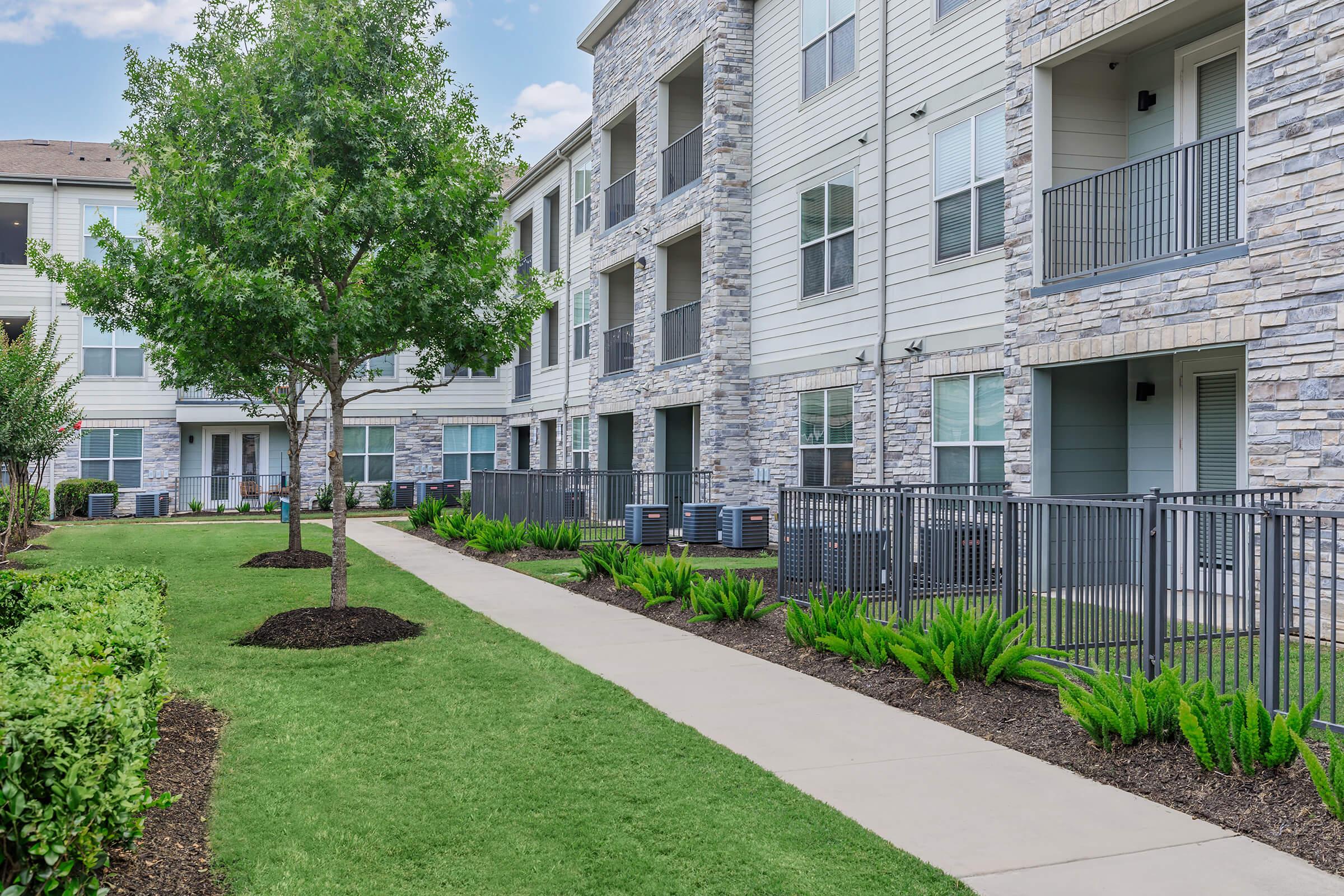 A landscaped pathway runs through a residential area featuring modern apartment buildings with stone accents. The greenery includes shrubs and a tree, while fences surround outdoor units leading to balconies. The scene conveys a clean, well-maintained living environment.