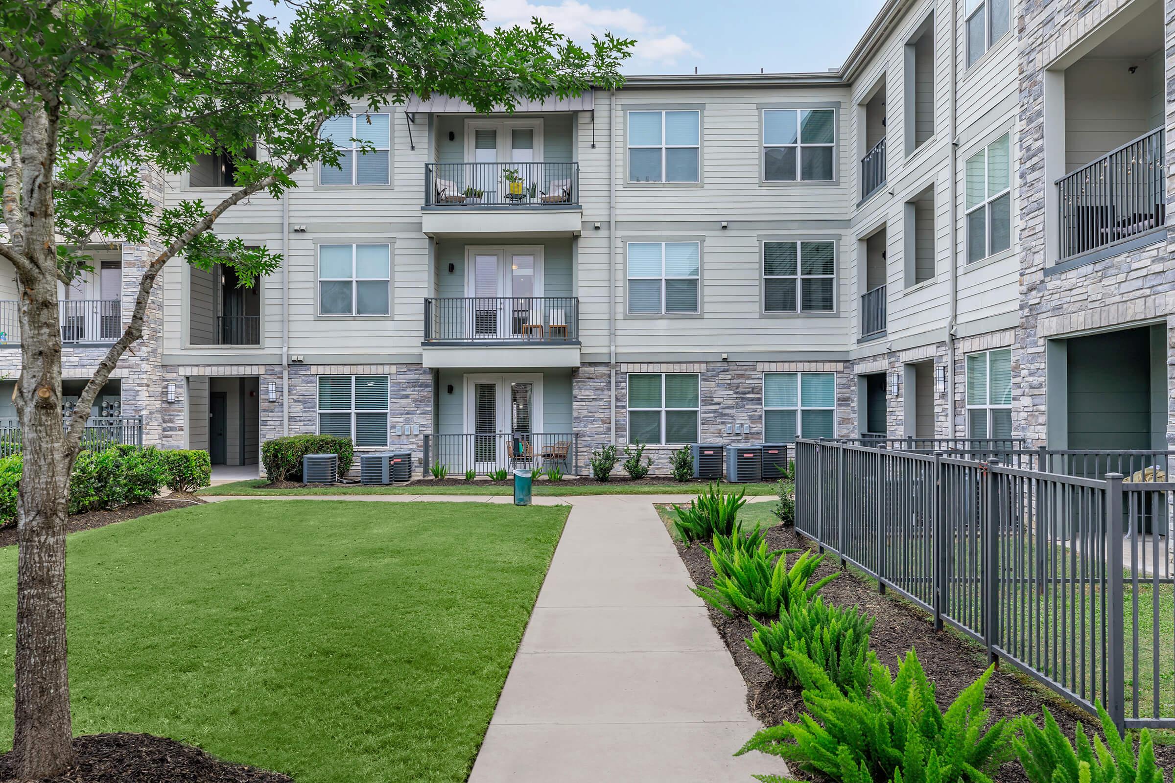 A well-maintained apartment complex with a grassy area and neatly trimmed hedges. The building features multiple balconies, large windows, and a stone facade. A paved walkway runs through the landscape, with air conditioning units visible near the ground level.