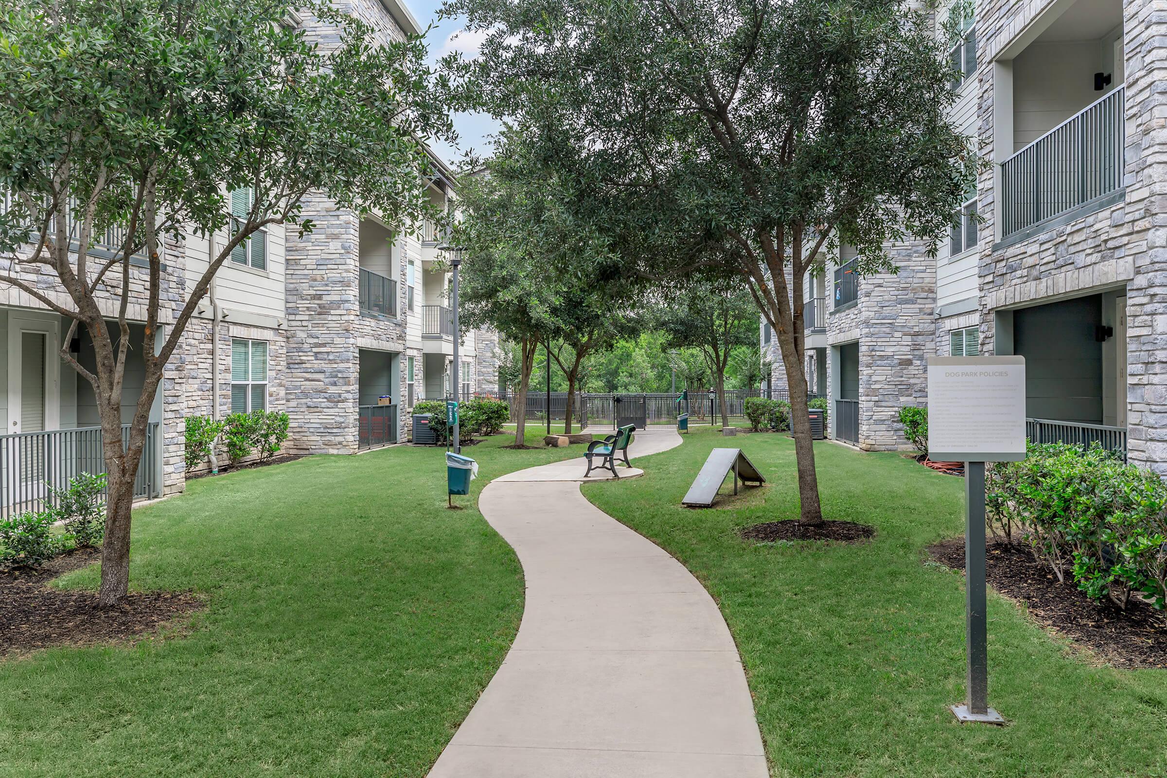 Pathway winding through a landscaped courtyard with greenery, benches, and a sign. The buildings have stone accents and balconies, creating a serene environment. Sunlight filters through the trees, enhancing the inviting atmosphere.