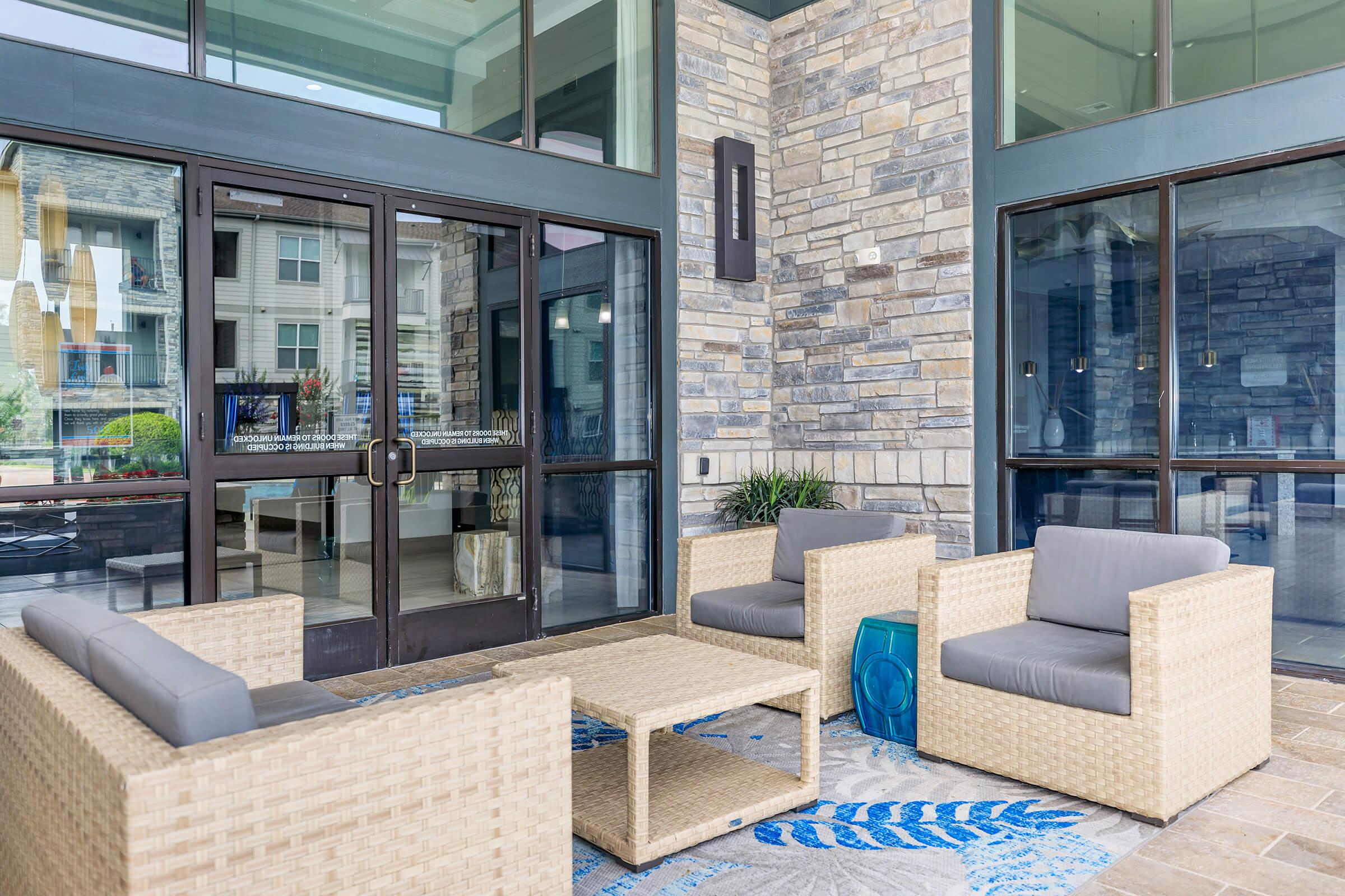 Seating area with wicker furniture, including two chairs and a coffee table, in a modern lobby. Large glass doors lead outside, showcasing a well-lit entrance. The floor features a blue and white patterned rug, and the walls are made of polished stone, creating a stylish and inviting atmosphere.