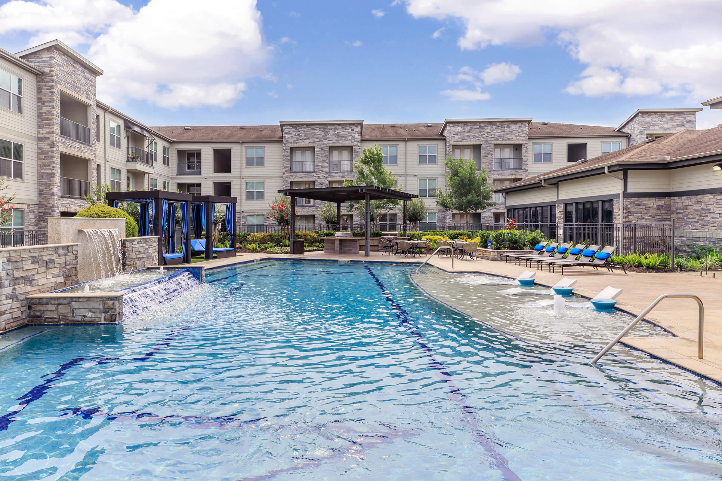A spacious outdoor pool area surrounded by modern apartment buildings. The pool features a waterfall, lounge chairs, and a shaded cabana. Lush landscaping adds to the serene atmosphere, making it an inviting space for relaxation and leisure. The sky is bright with a few clouds.