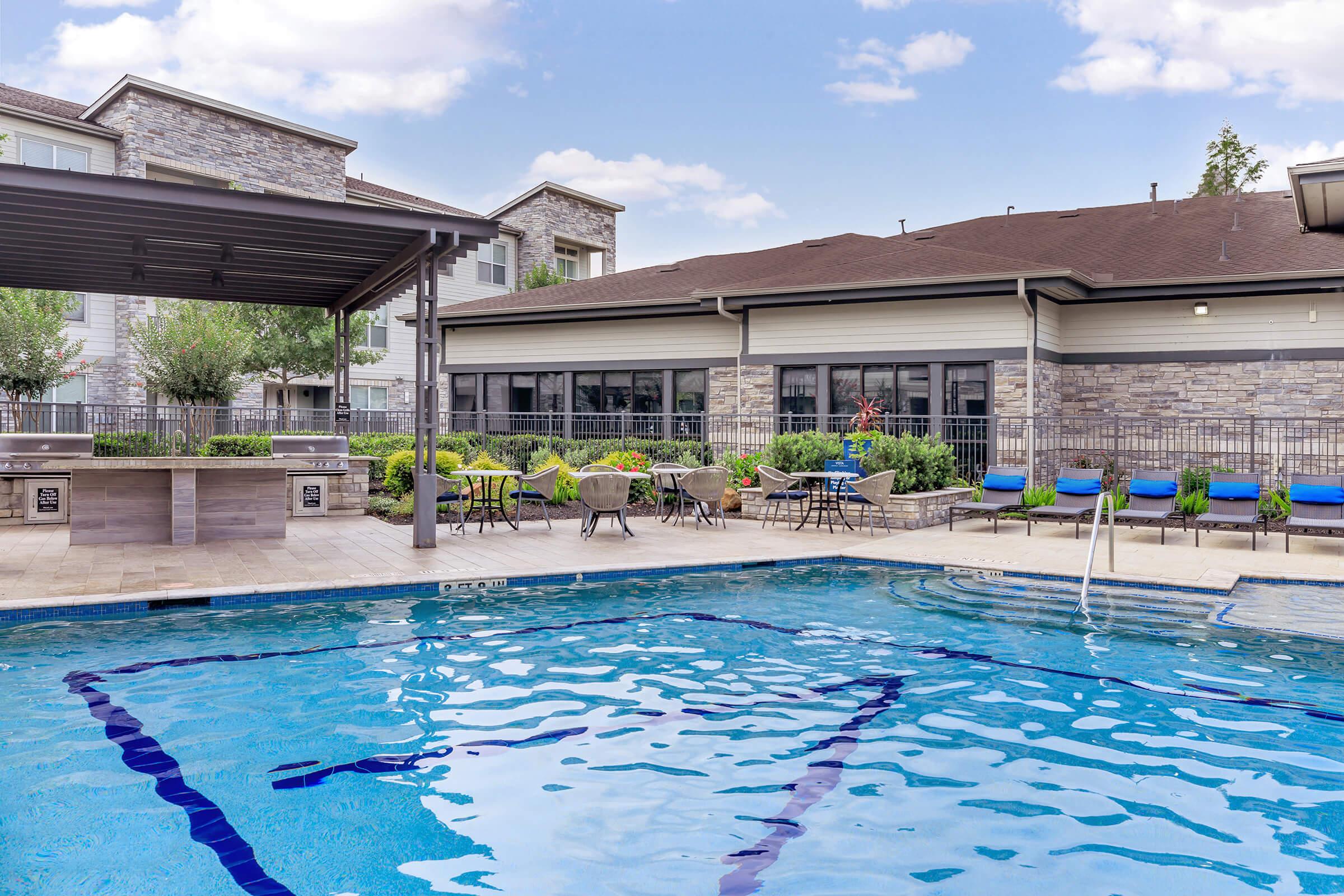 A sparkling outdoor swimming pool surrounded by lounge chairs and tables. The area includes a shaded seating space with modern furniture, greenery, and a well-maintained stone building in the background, offering a comfortable and inviting atmosphere for relaxation and leisure.