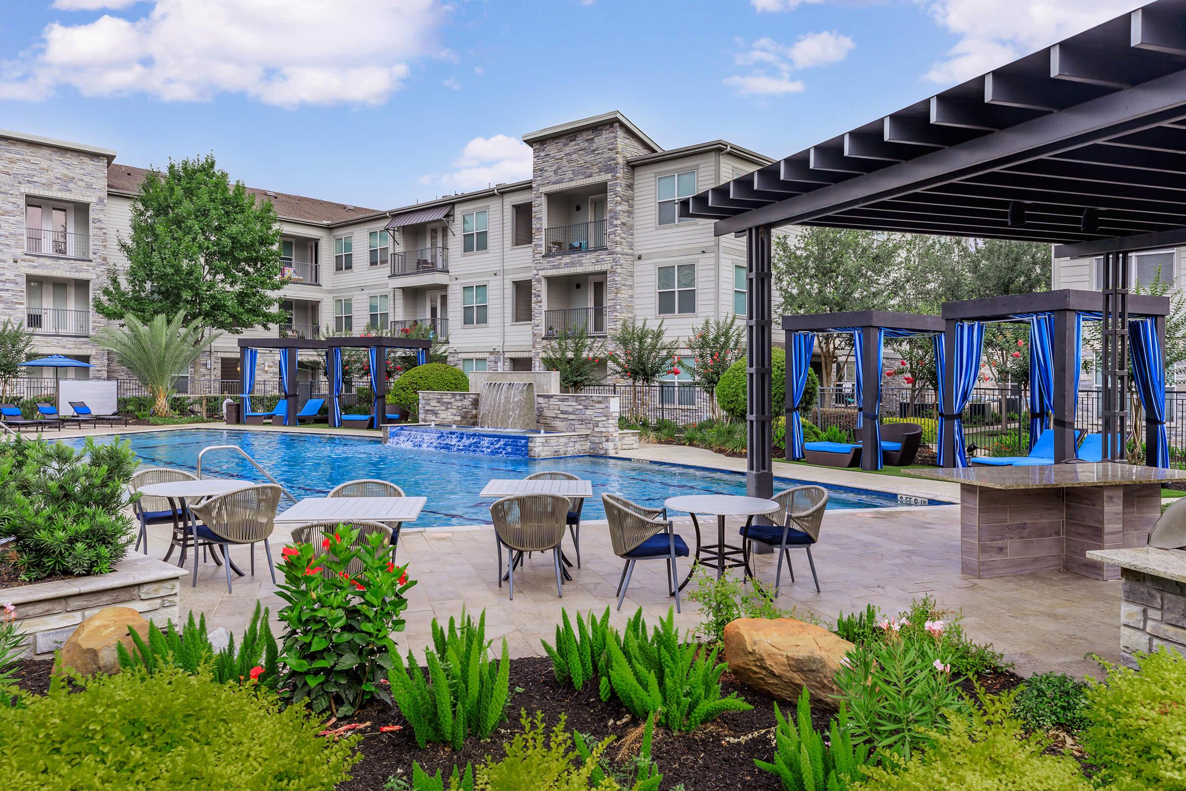 A well-maintained apartment complex pool area featuring a crystal-clear pool surrounded by lounge chairs and cabanas. Lush greenery and flowers add color to the landscape, while modern buildings provide a stylish backdrop. The sky is partly cloudy, creating a pleasant atmosphere for relaxation.