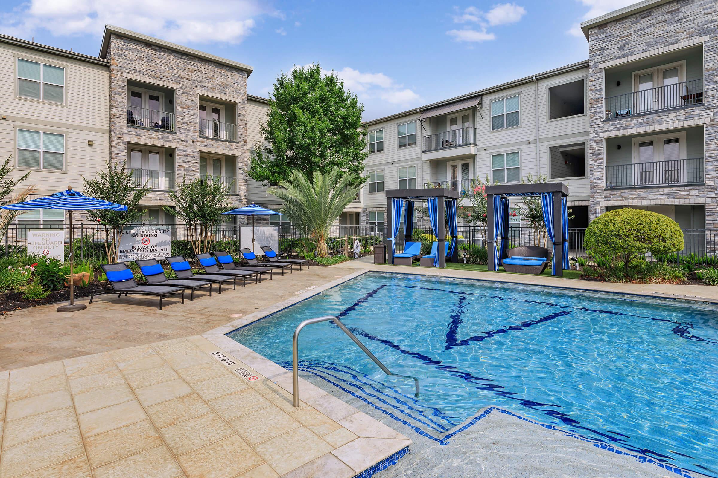 A well-maintained outdoor pool area surrounded by lounge chairs and blue umbrellas, with two cabanas nearby. Modern apartment buildings are visible in the background, set against a clear blue sky with a few clouds. Lush greenery adds to the inviting atmosphere of the space.