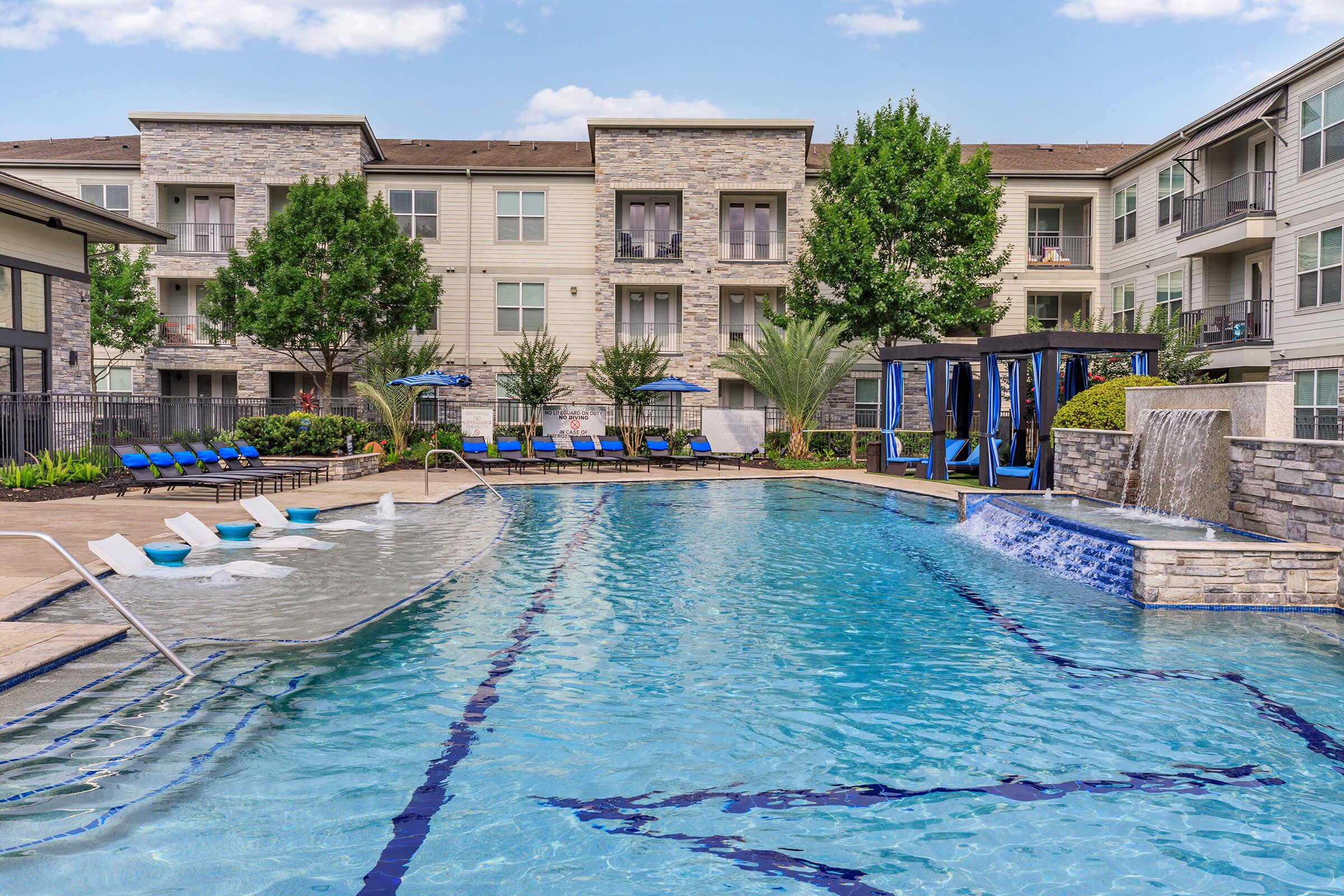 A luxurious apartment complex pool area featuring a spacious swimming pool with a waterfall, surrounded by lounge chairs and lush greenery. The modern buildings in the background add to the ambiance, creating a relaxing outdoor space for residents.